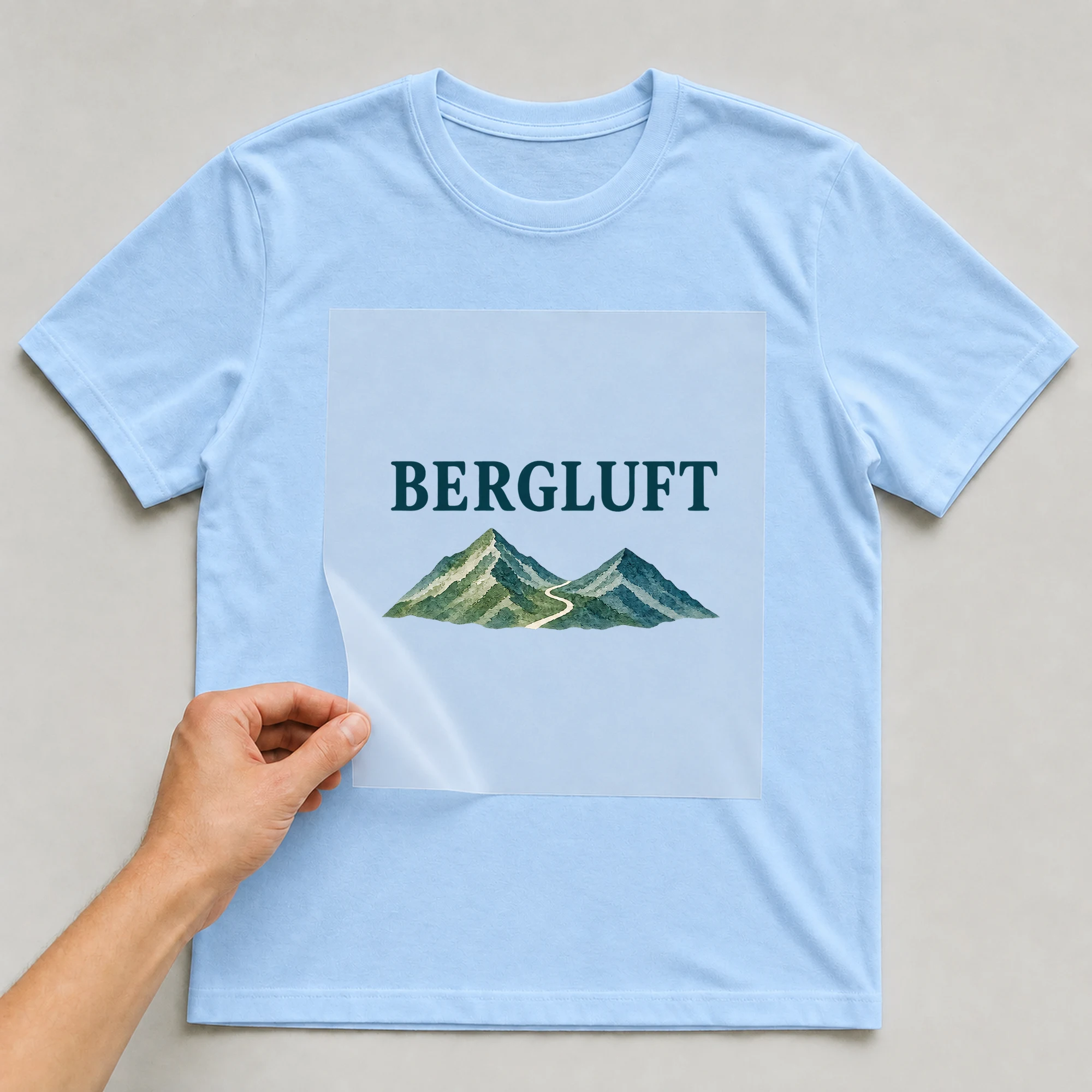 Bügelbild „BERGLUFT“ auf babyblauem T-Shirt, Folie wird aufgelegt