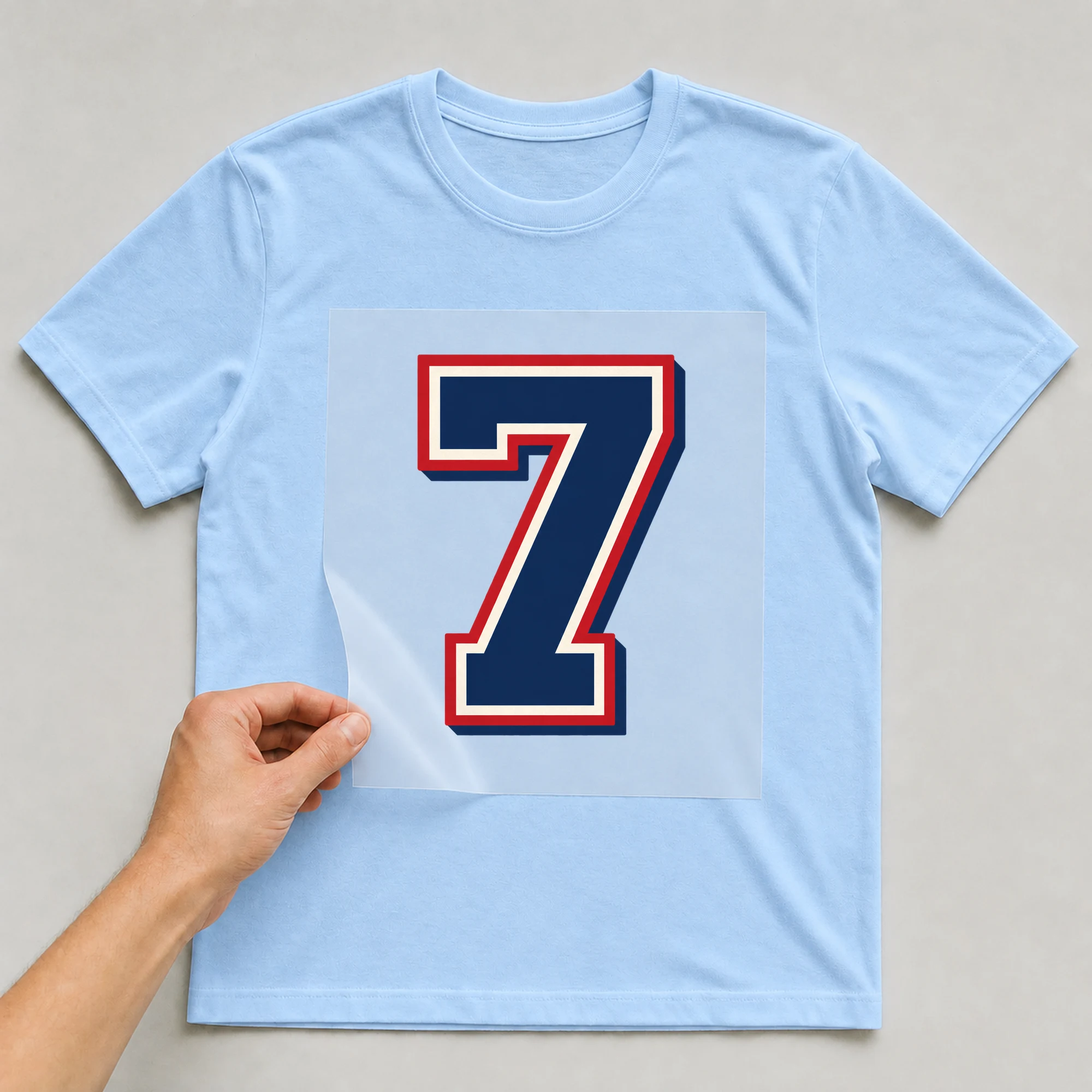 Bügelbild „Zahl 7 im Retro-College-Stil“ auf babyblauem T-Shirt, Folie wird aufgelegt