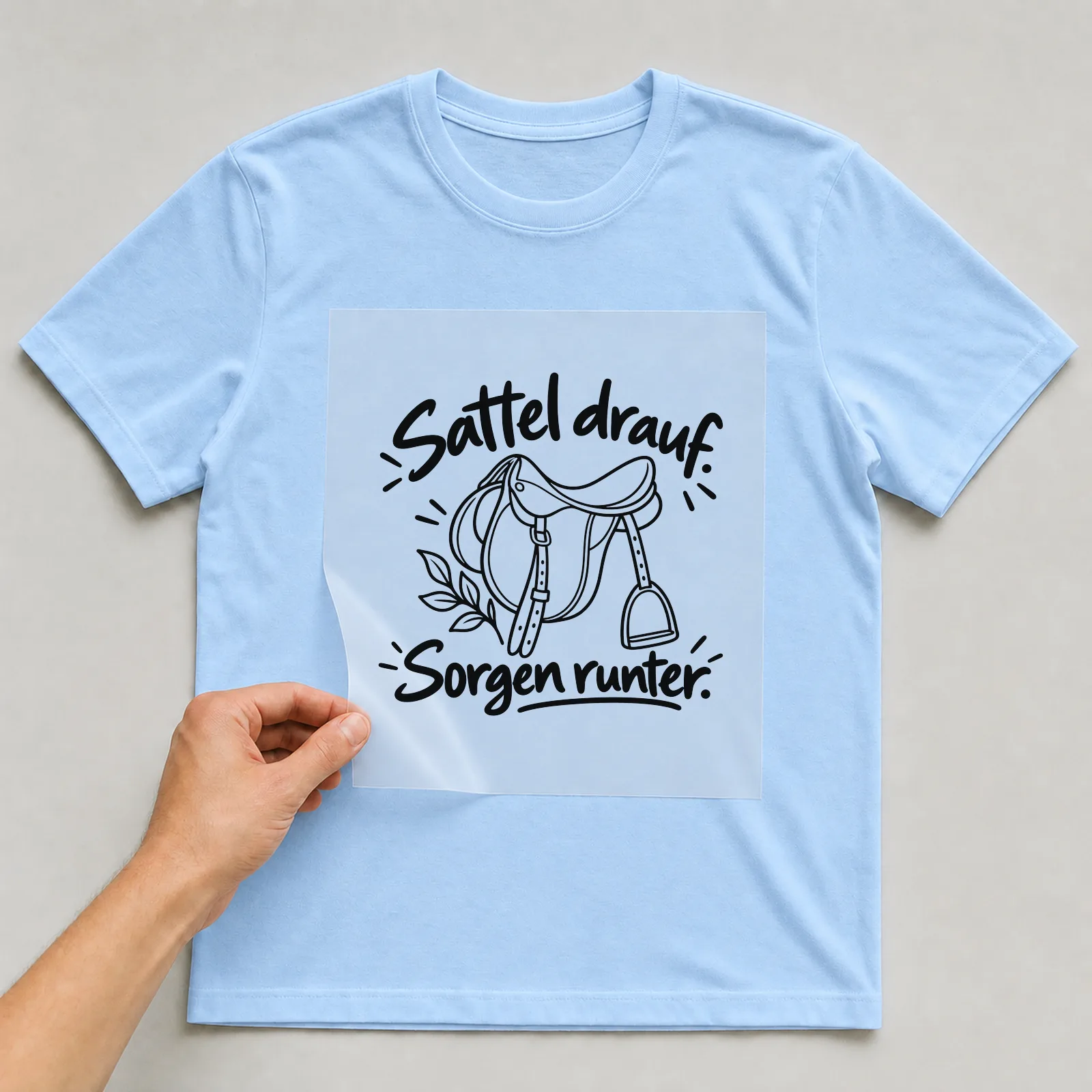 Bügelbild „Sattel drauf. Sorgen runter.“ auf hellblauem T-Shirt, Folie wird aufgelegt