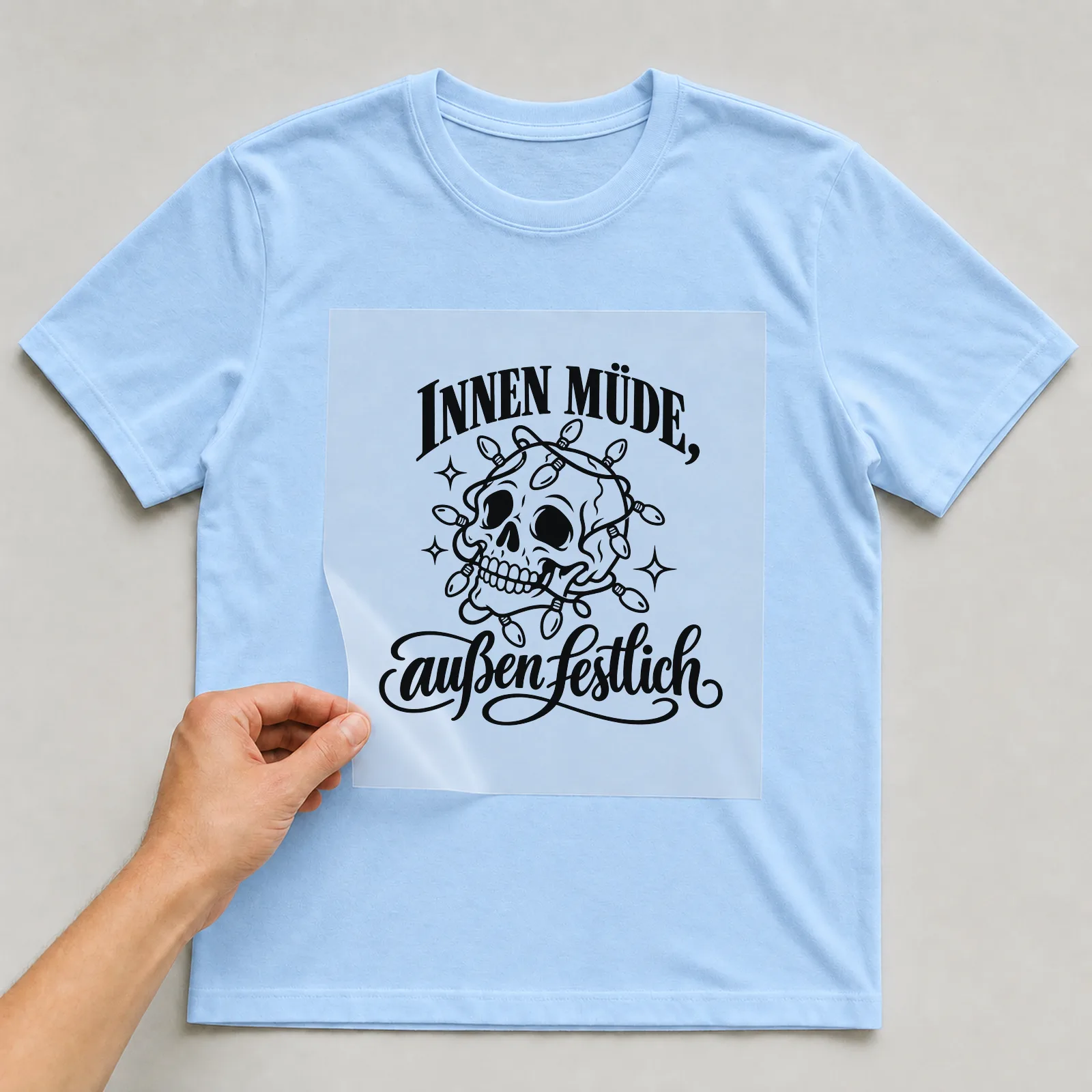 Bügelbild „Innen müde, außen festlich“ auf hellblauem T-Shirt, Folie wird aufgelegt
