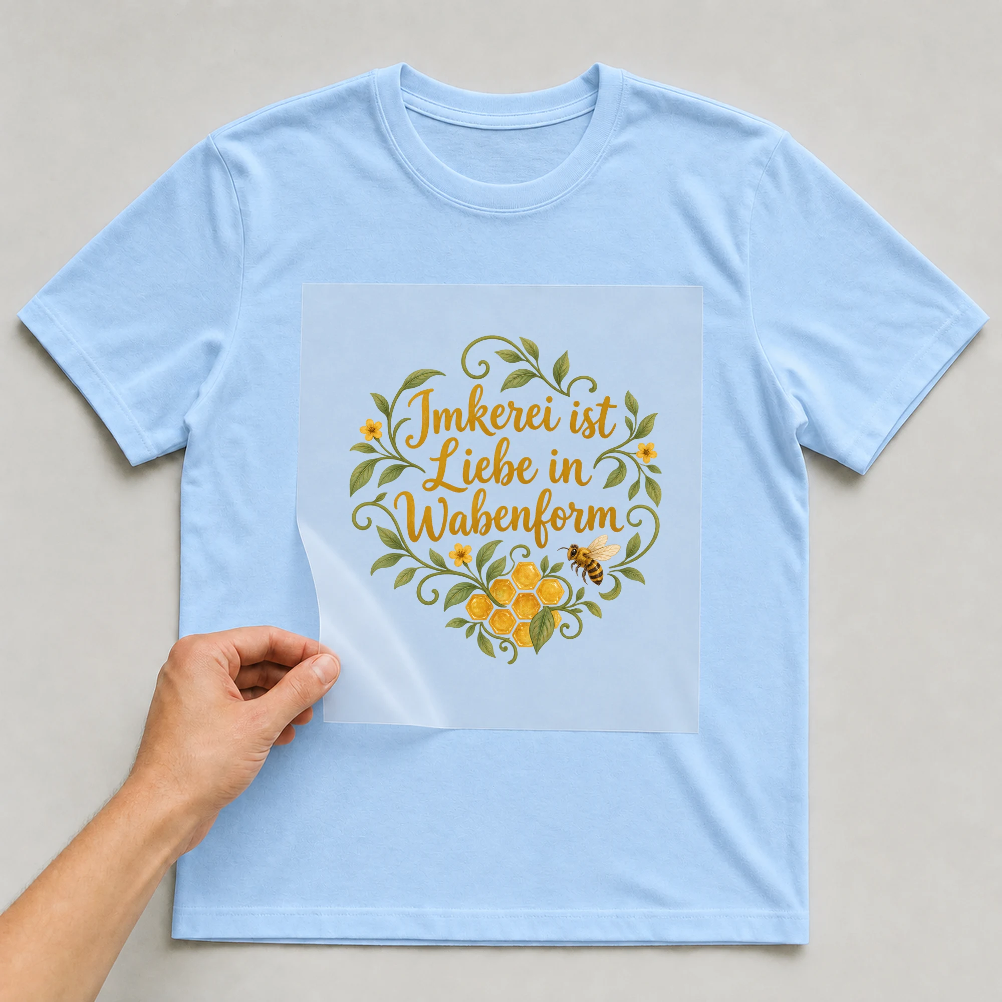 Bügelbild „Imkerei ist Liebe in Wabenform“ auf babyblauem T-Shirt, Folie wird aufgelegt
