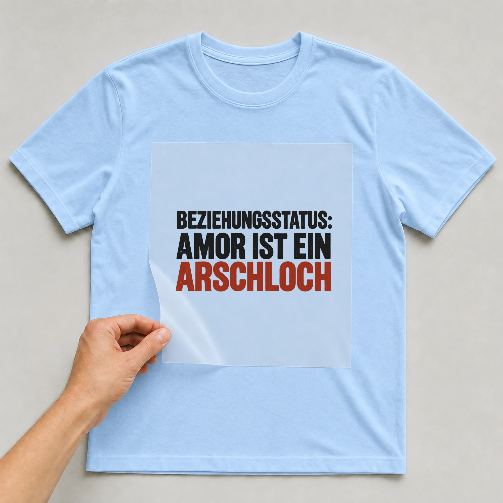 Bügelbild „Beziehungsstatus: Amor ist ein Ar*chloch“ auf babyblauem T-Shirt, Folie wird aufgelegt