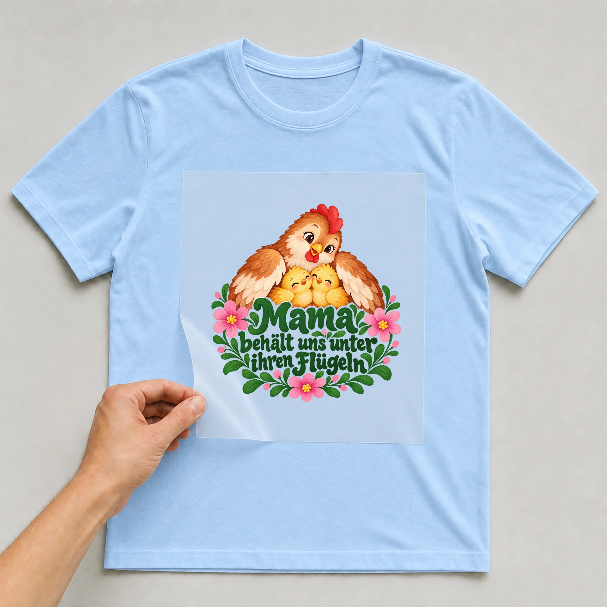 Bügelbild „Mama behält uns unter ihren Flügeln“ auf hellblauem T-Shirt, Folie wird aufgelegt