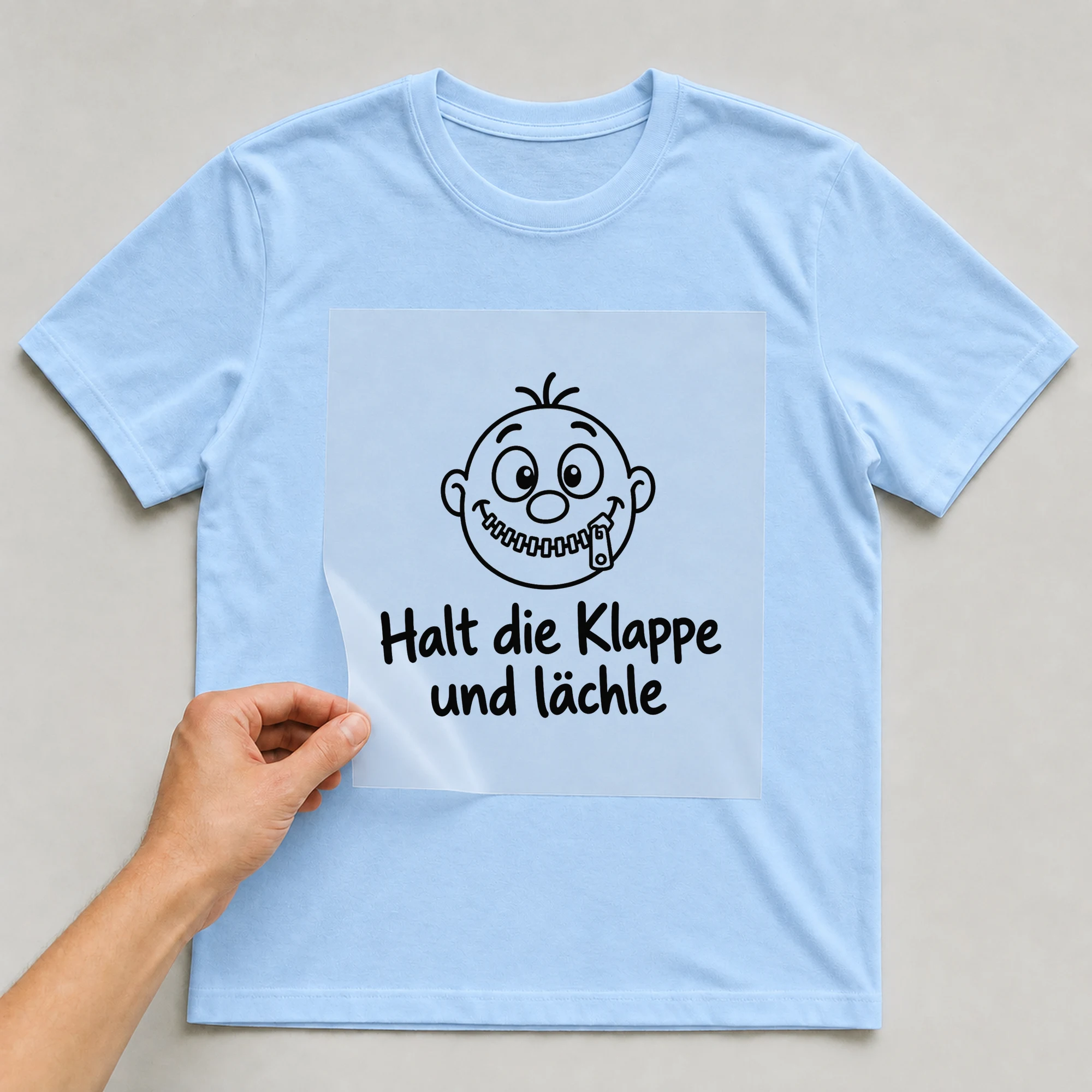 Bügelbild „Halt die Klappe und lächle“ auf babyblauem T-Shirt, Folie wird aufgelegt