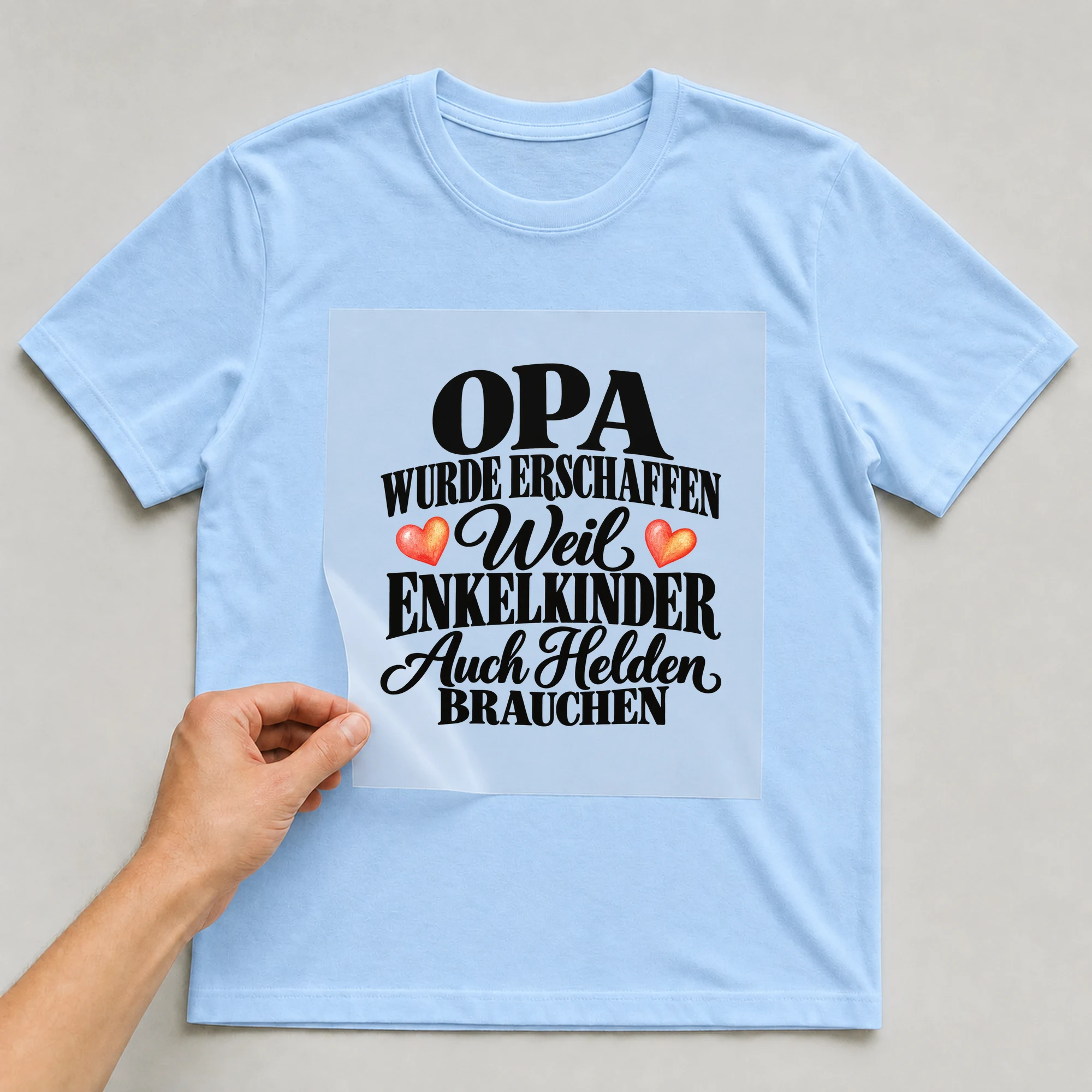 Bügelbild „Opa wurde erschaffen weil Enkelkinder auch Helden brauchen“ auf babyblauem T-Shirt, Folie wird aufgelegt