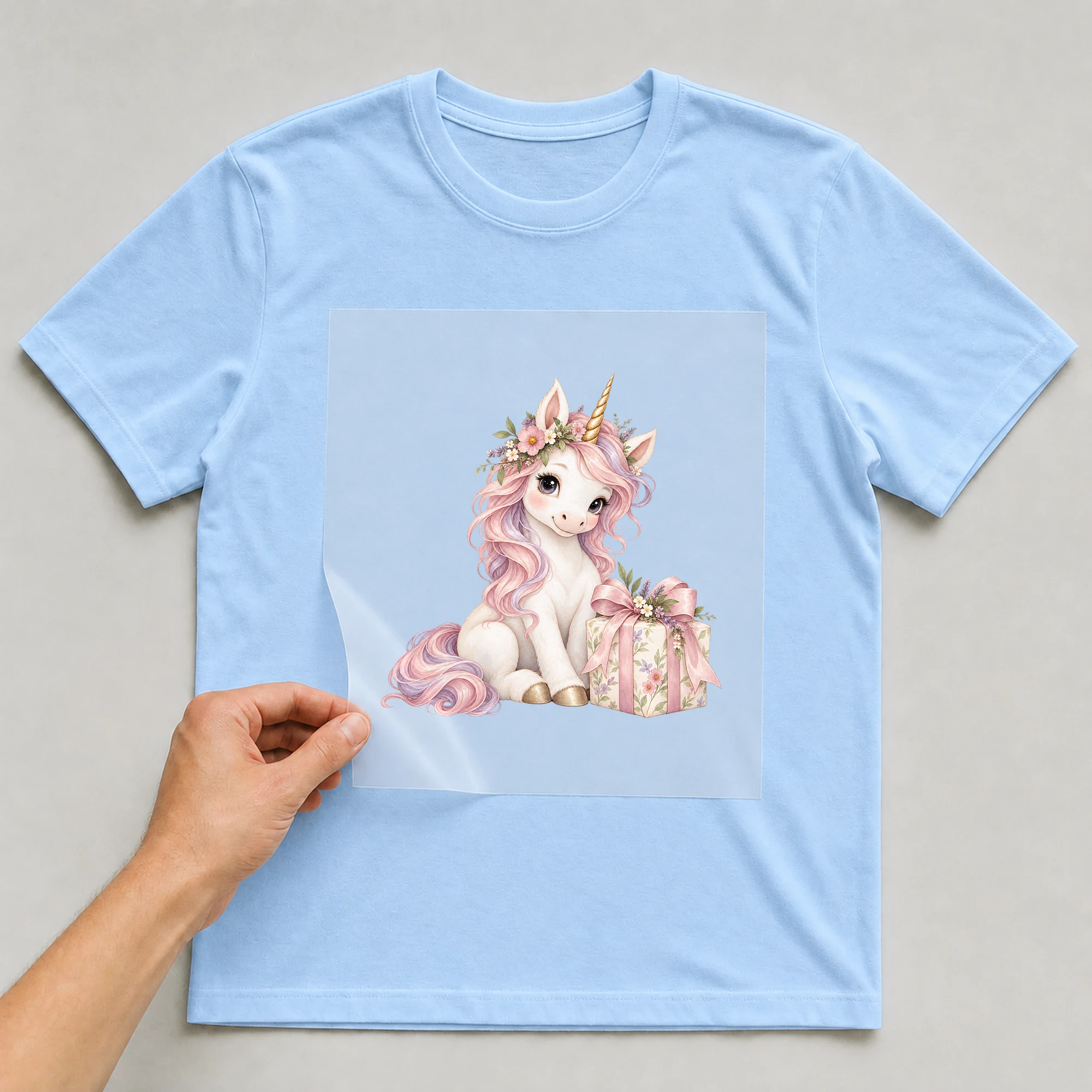 Bügelbild „Einhorn mit Blumen“ auf babyblauem T-Shirt, Folie wird aufgelegt