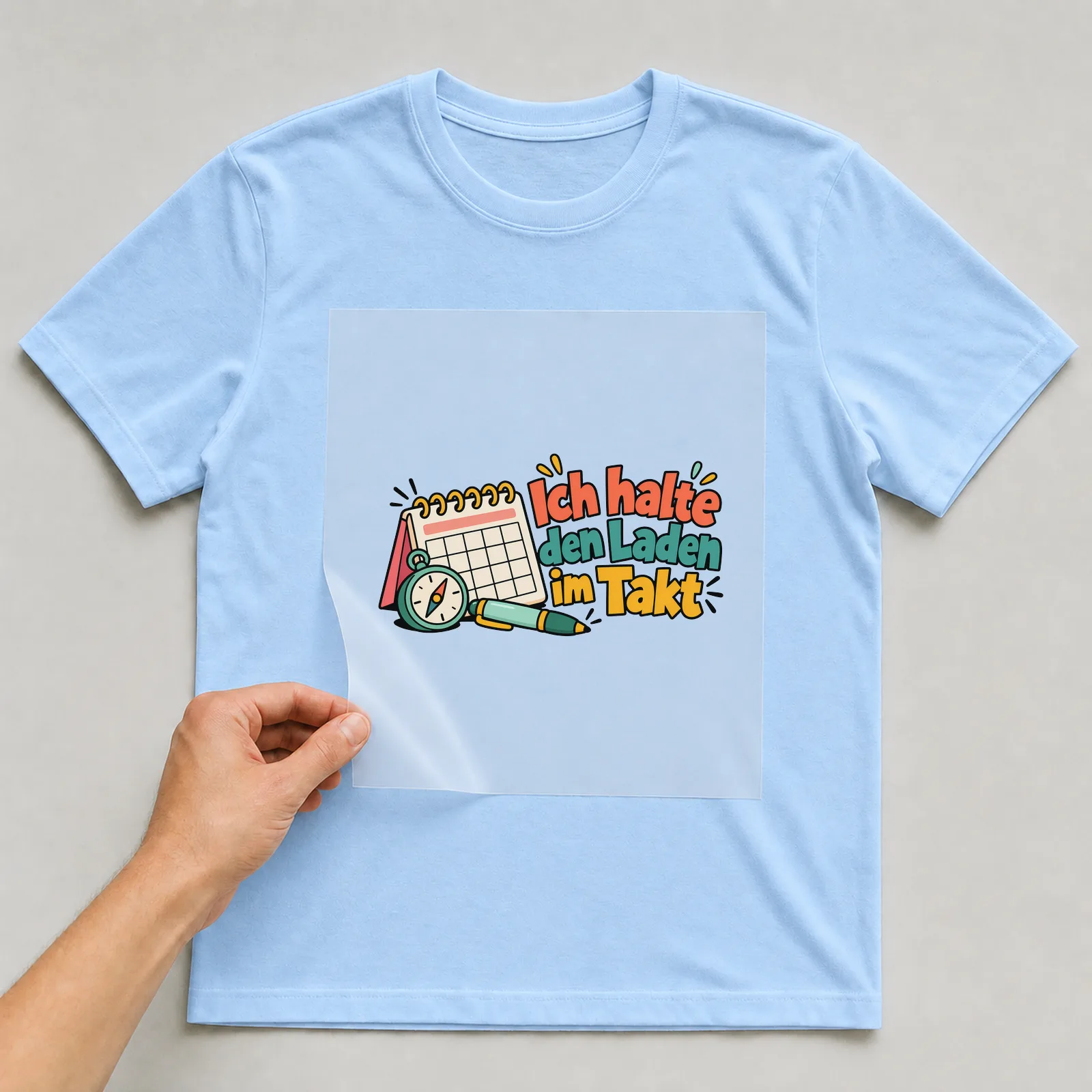 Bügelbild „Ich halte den Laden im Takt“ auf hellblauem T-Shirt, Folie wird aufgelegt