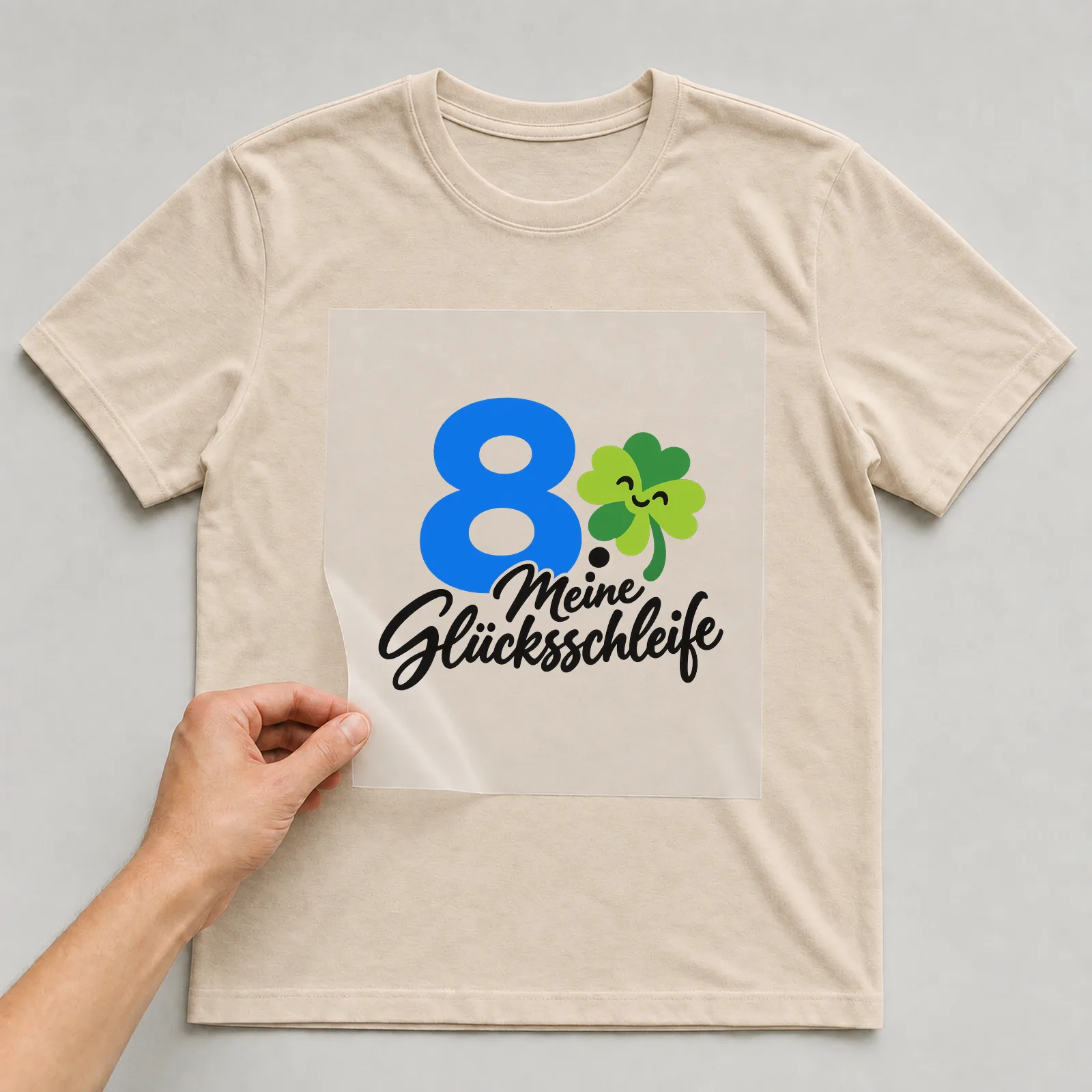 Bügelbild „8 · Meine Glücksschleife“ auf beigem T-Shirt, Folie wird aufgelegt