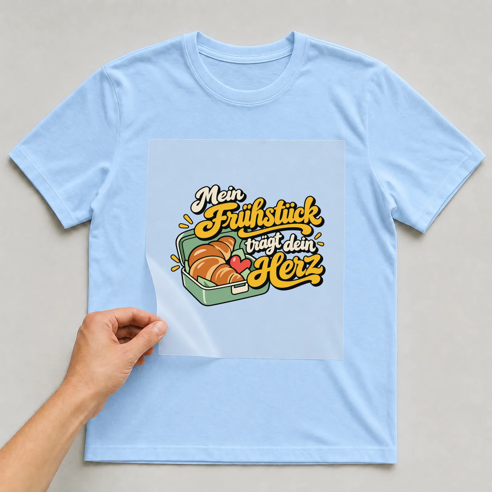 Bügelbild „Mein Frühstück trägt dein Herz“ auf hellblauem T-Shirt, Folie wird aufgelegt