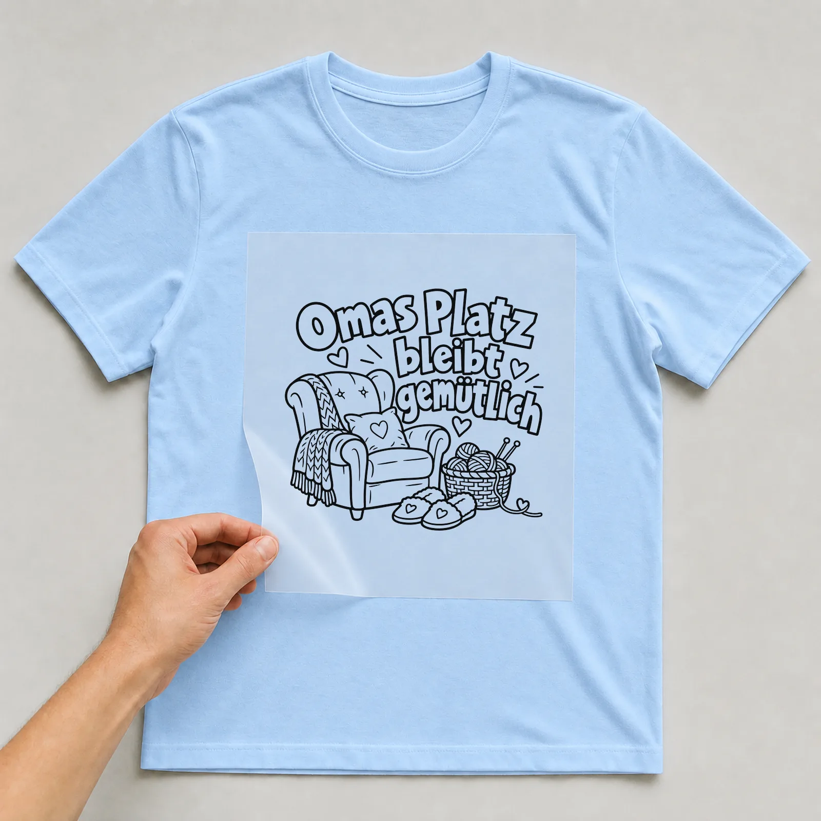 Bügelbild „Omas Platz bleibt gemütlich“ auf hellblauem T-Shirt, Folie wird aufgelegt