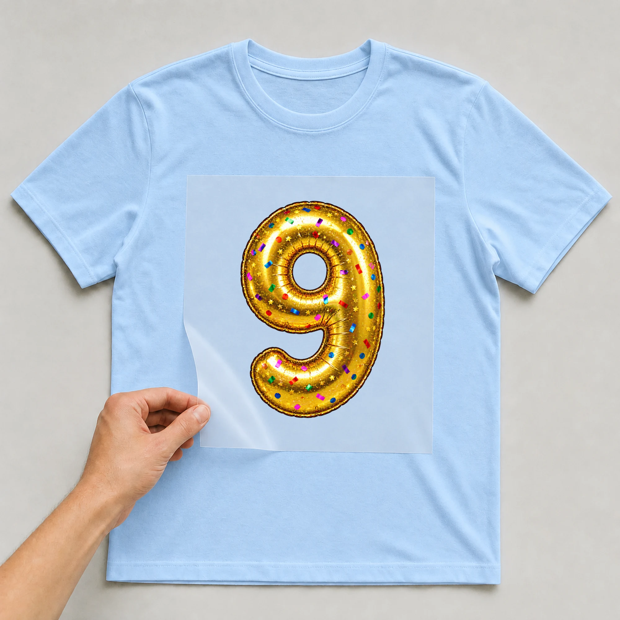Bügelbild „Zahl 9 im Konfetti-Metallic-Look“ auf babyblauem T-Shirt, Folie wird aufgelegt