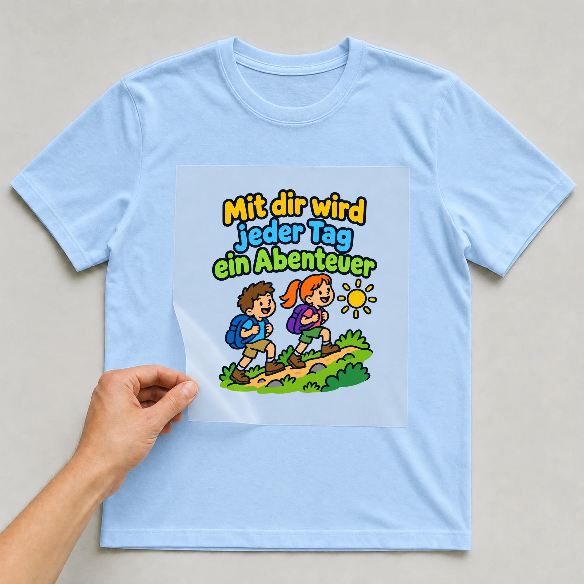 Bügelbild „Mit dir wird jeder Tag ein Abenteuer“ auf babyblauem T-Shirt, Folie wird aufgelegt
