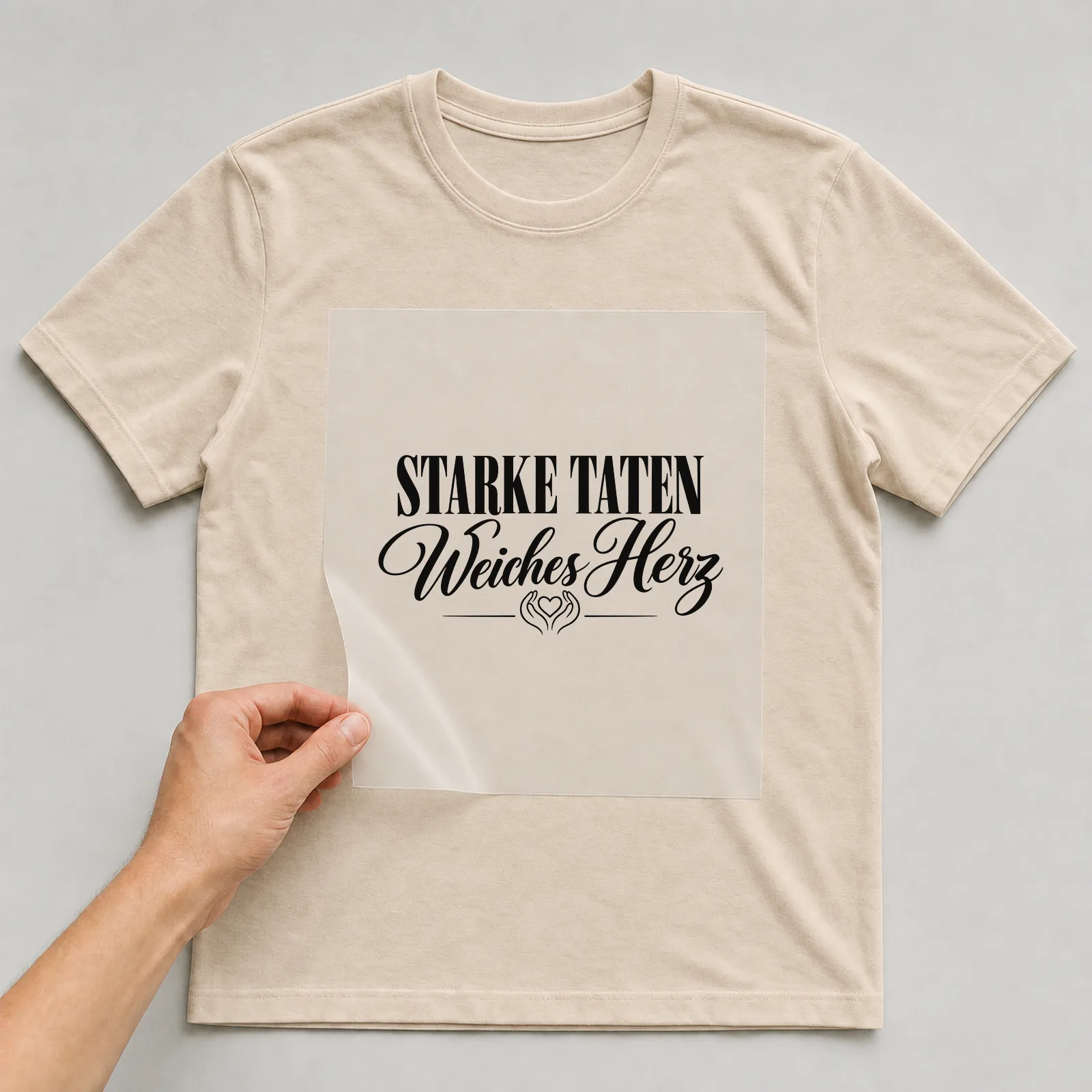 Bügelbild „Starke Taten Weiches Herz“ auf beigem T-Shirt, Folie wird aufgelegt