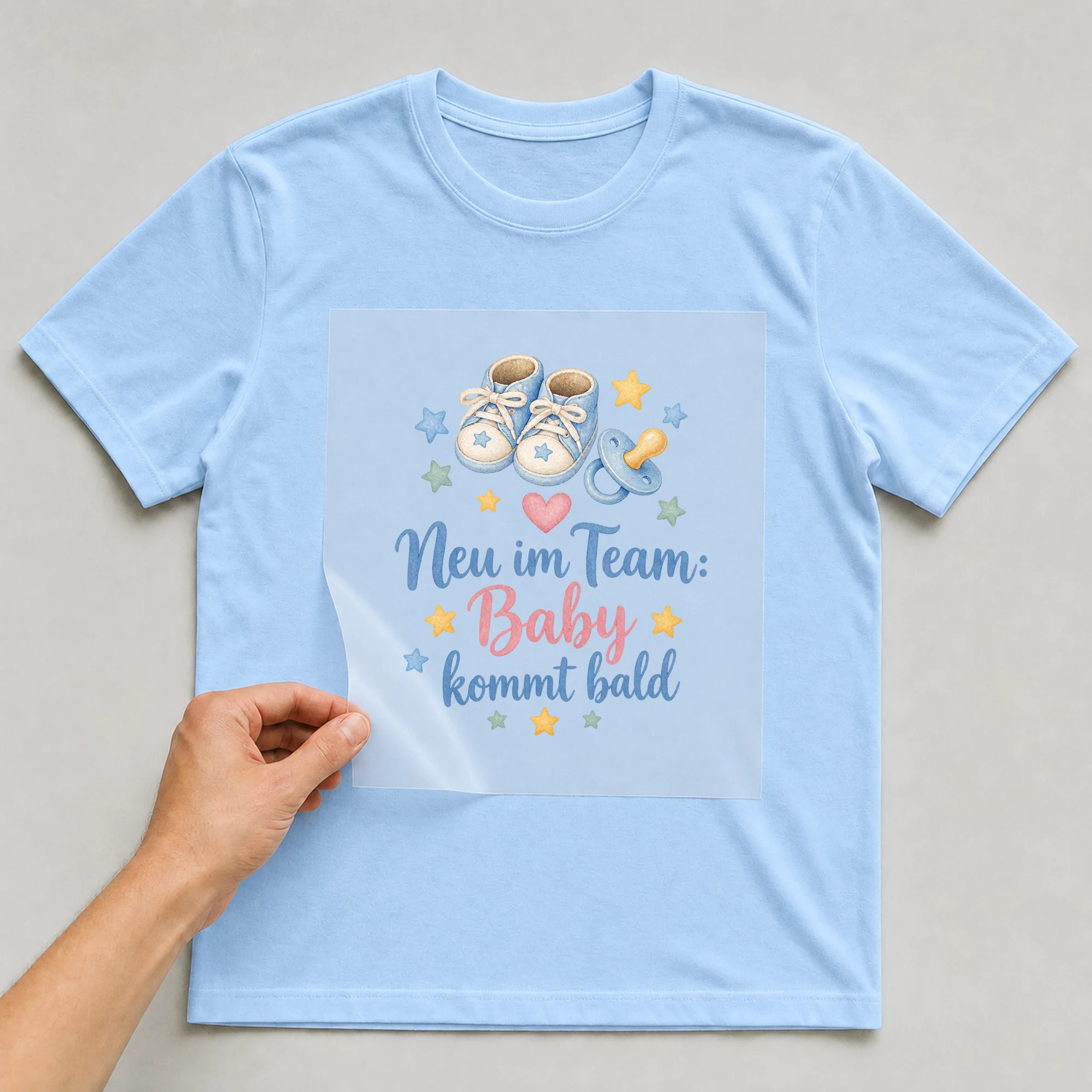 Bügelbild „Neu im Team: Baby kommt bald“ auf babyblauem T-Shirt, Folie wird aufgelegt