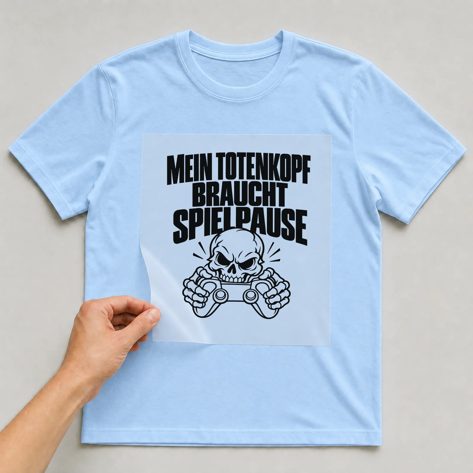 Bügelbild „Mein Totenkopf braucht Spielpause“ auf hellblauem T-Shirt, Folie wird aufgelegt