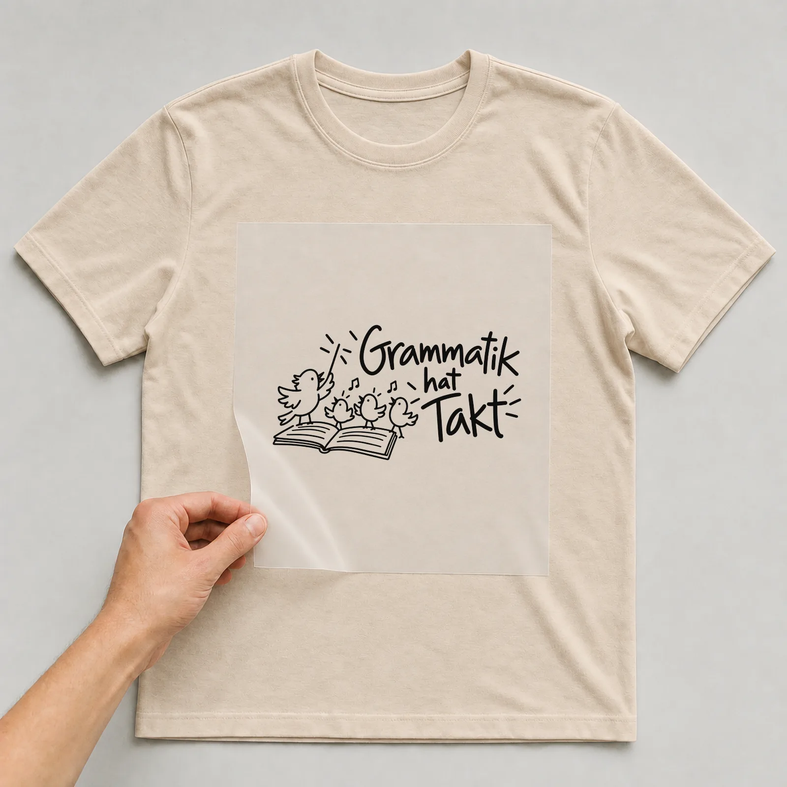 Bügelbild „Grammatik hat Takt“ auf beigem T-Shirt, Folie wird aufgelegt