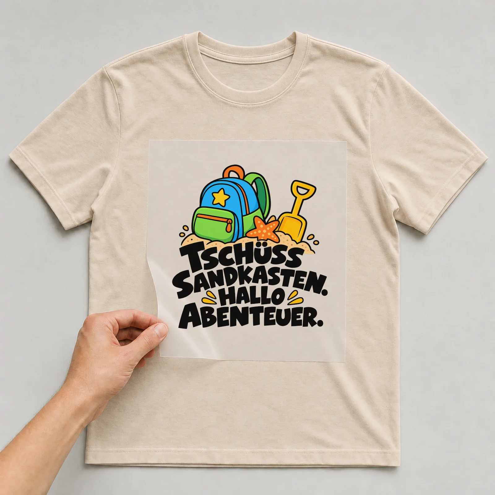 Bügelbild „Tschüss Sandkasten. Hallo Abenteuer.“ auf beigem T-Shirt, Folie wird aufgelegt