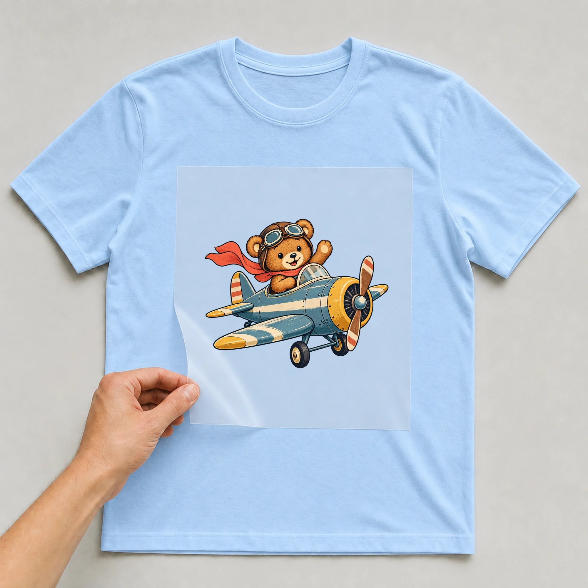 Bügelbild „Flugzeug mit Bär“ auf babyblauem T-Shirt, Folie wird aufgelegt