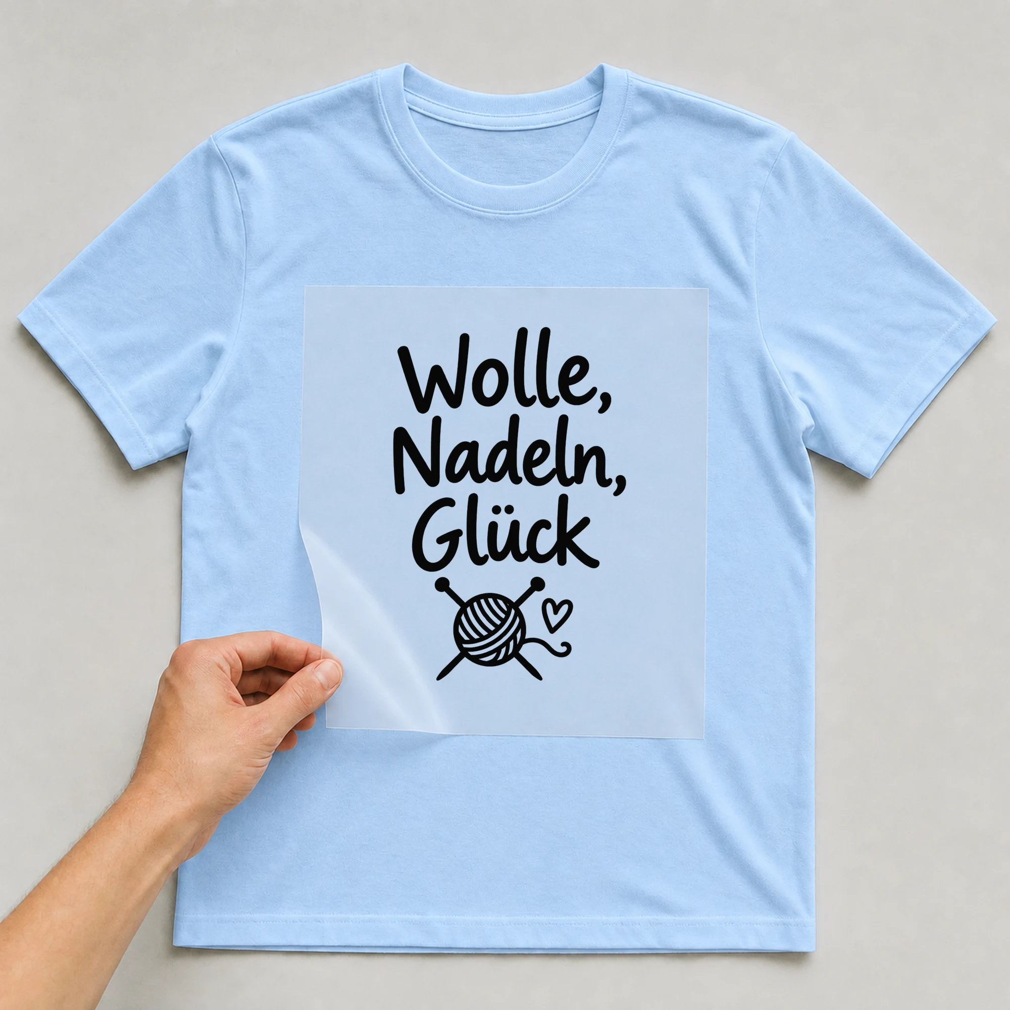 Bügelbild „Wolle, Nadeln, Glück“ auf babyblauem T-Shirt, Folie wird aufgelegt