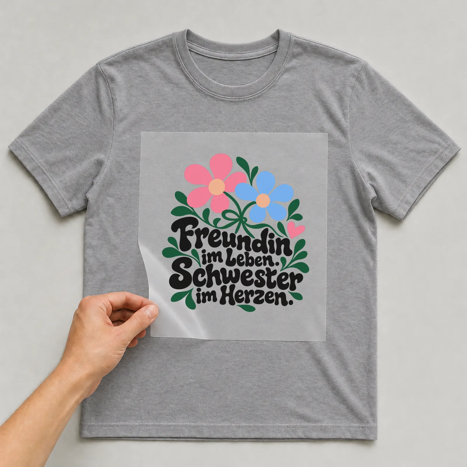 Bügelbild „Freundin im Leben. Schwester im Herzen.“ auf grauem T-Shirt, Folie wird aufgelegt