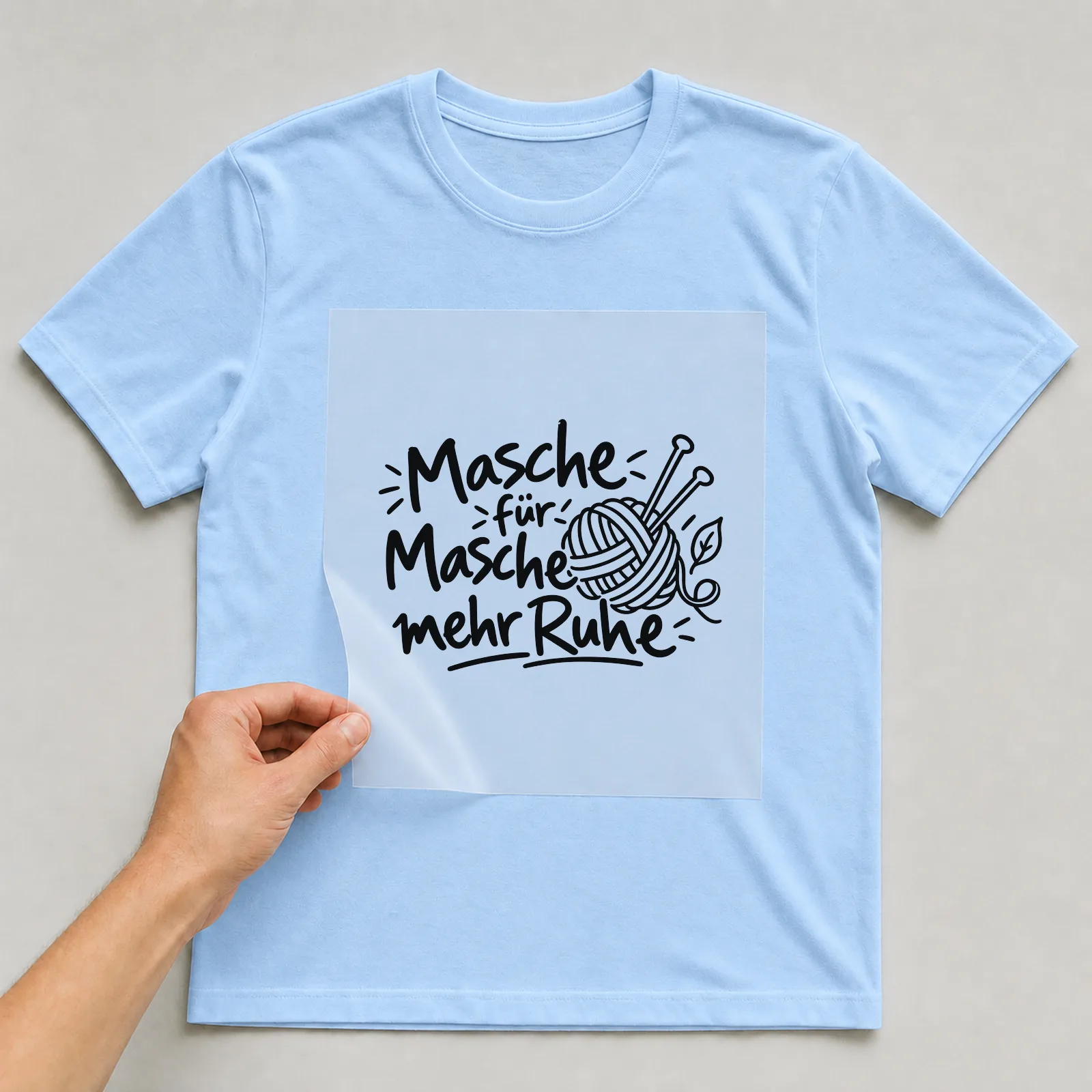 Bügelbild „Masche für Masche mehr Ruhe“ auf hellblauem T-Shirt, Folie wird aufgelegt
