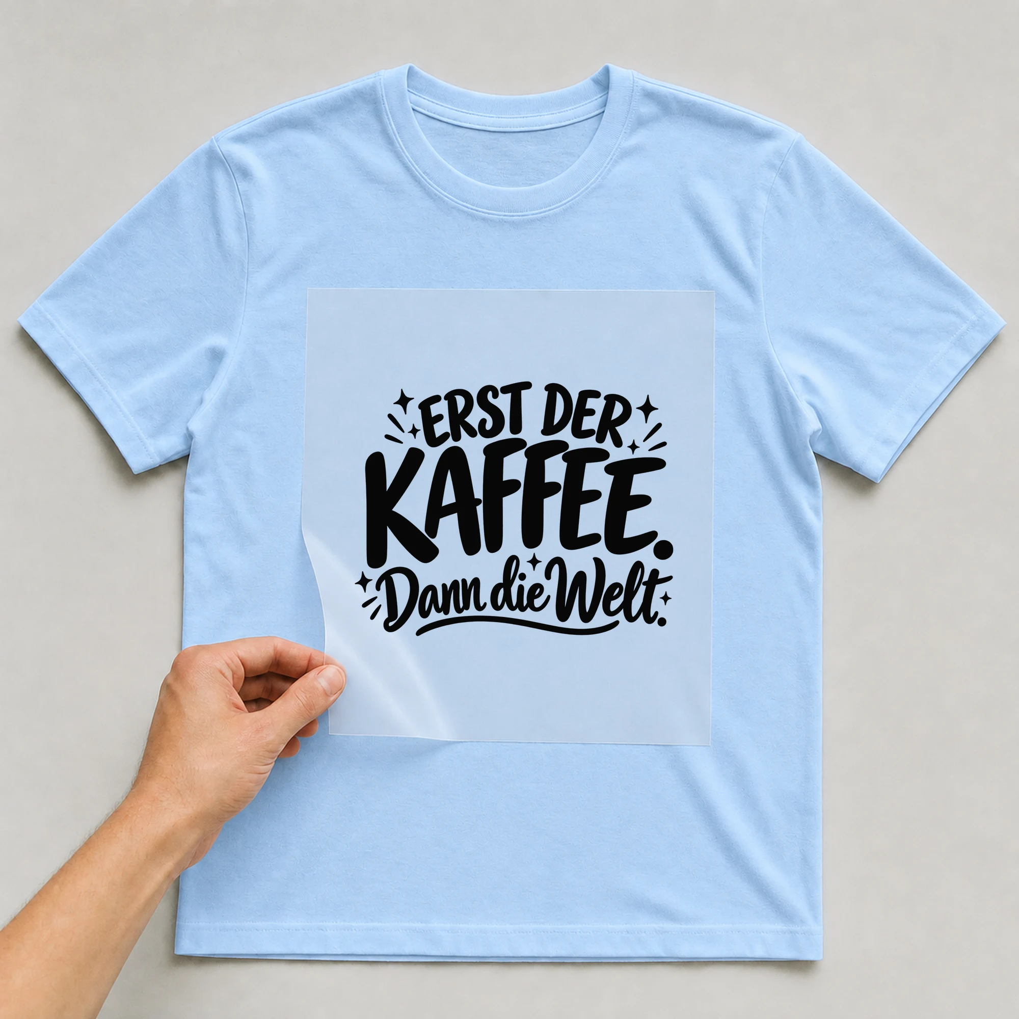 Bügelbild „Erst der Kaffee. Dann die Welt.“ auf babyblauem T-Shirt, Folie wird aufgelegt