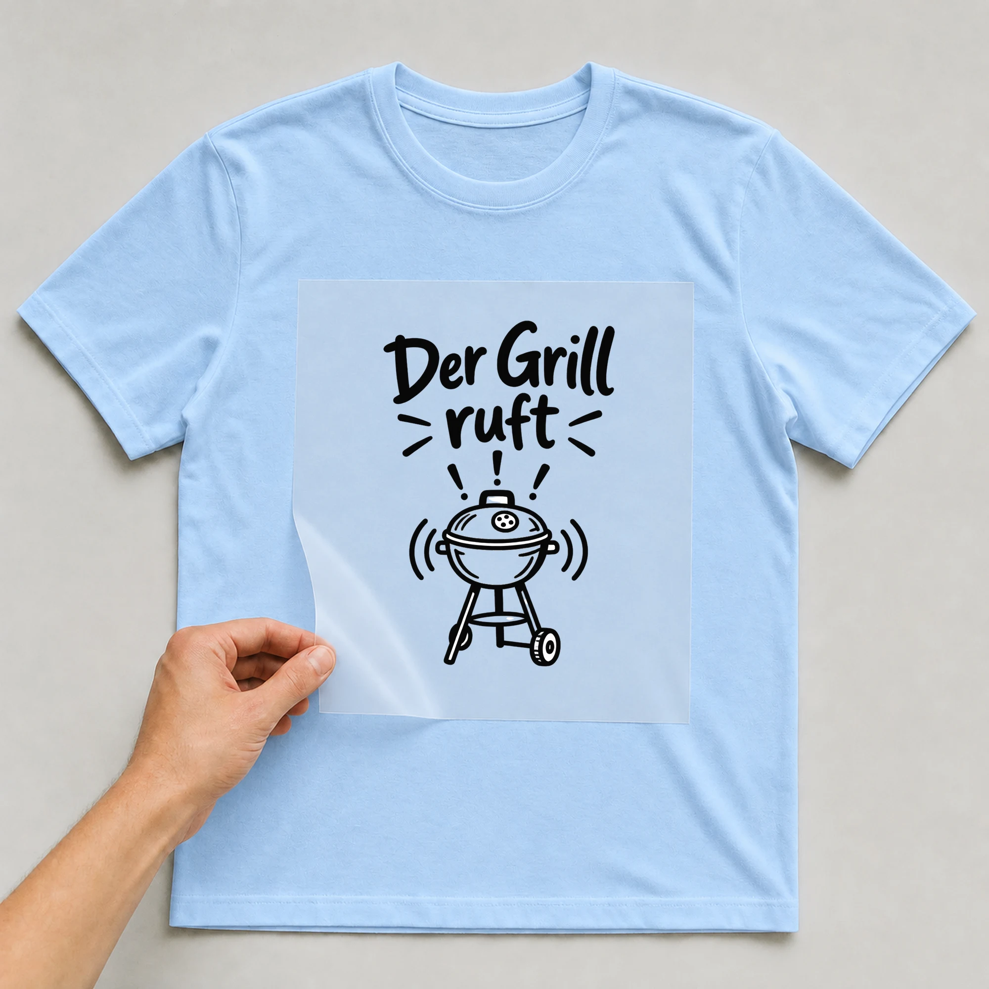 Bügelbild „Der Grill ruft“ auf babyblauem T-Shirt, Folie wird aufgelegt