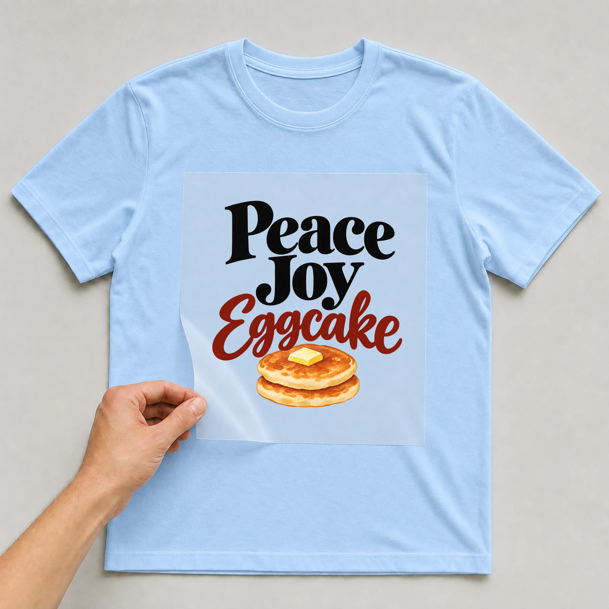 Bügelbild „Peace Joy Eggcake“ auf babyblauem T-Shirt, Folie wird aufgelegt