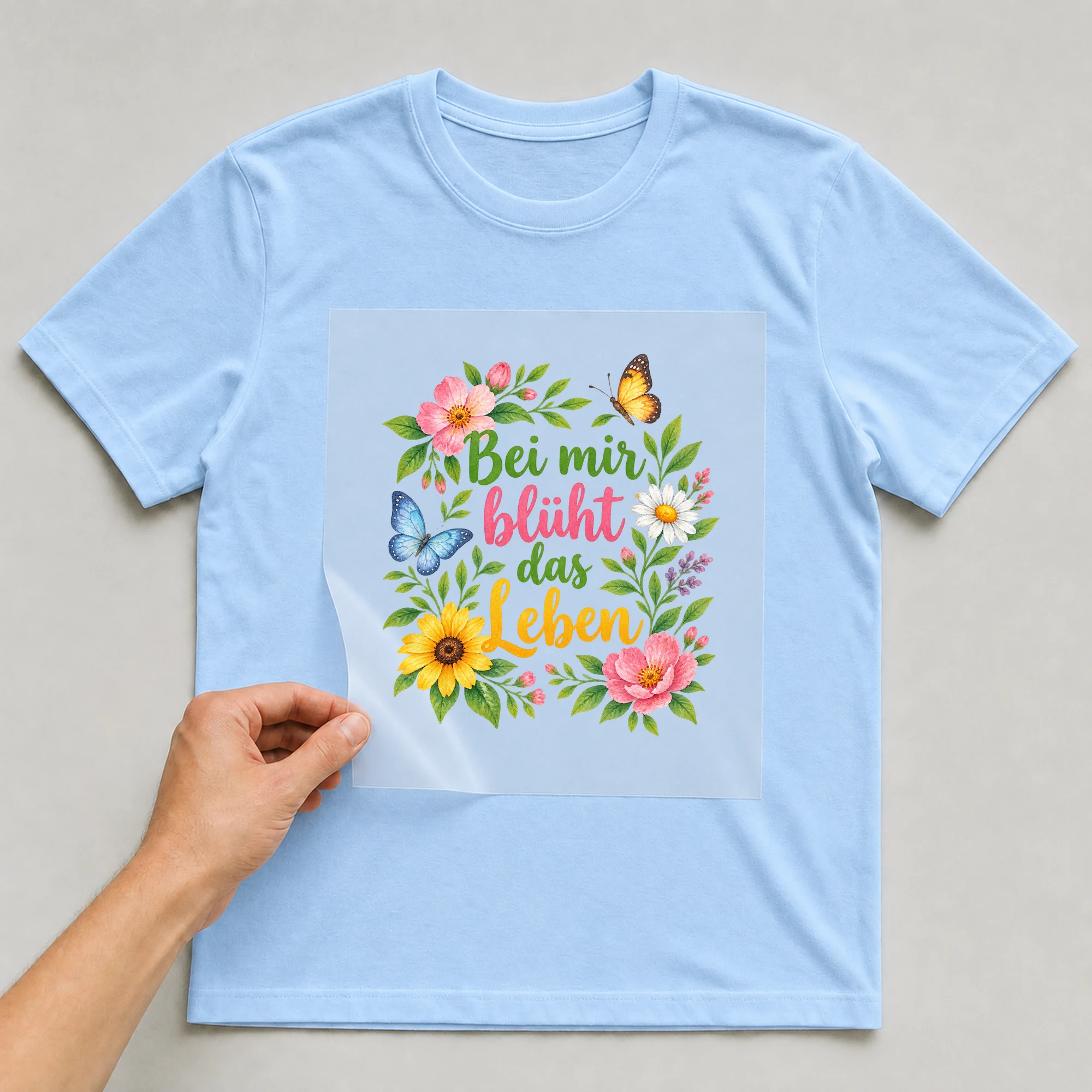 Bügelbild „Bei mir blueht das leben“ auf babyblauem T-Shirt, Folie wird aufgelegt