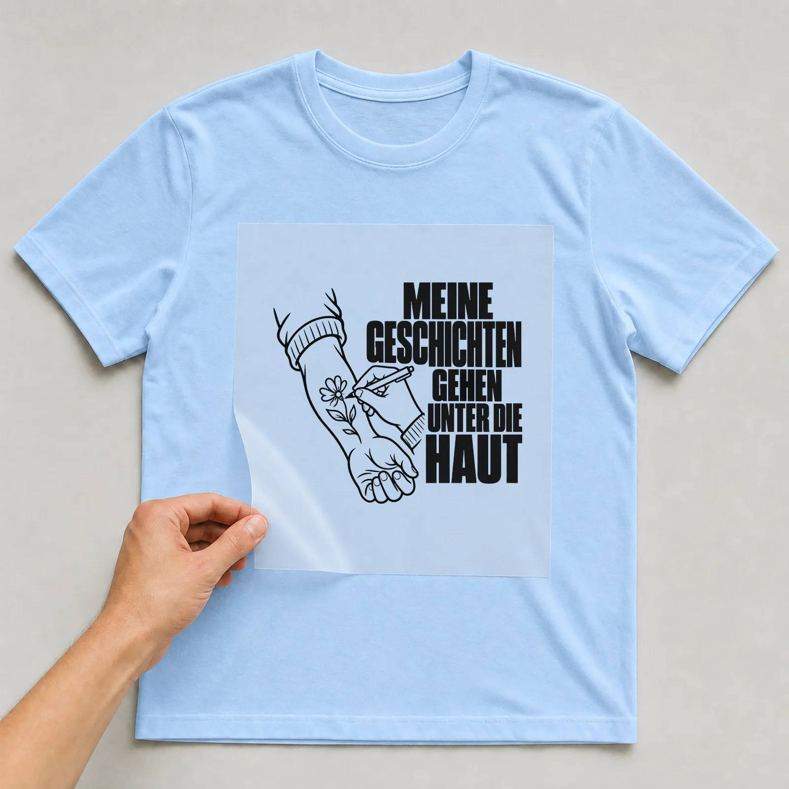 Bügelbild „Meine Geschichten gehen unter die Haut“ auf hellblauem T-Shirt, Folie wird aufgelegt