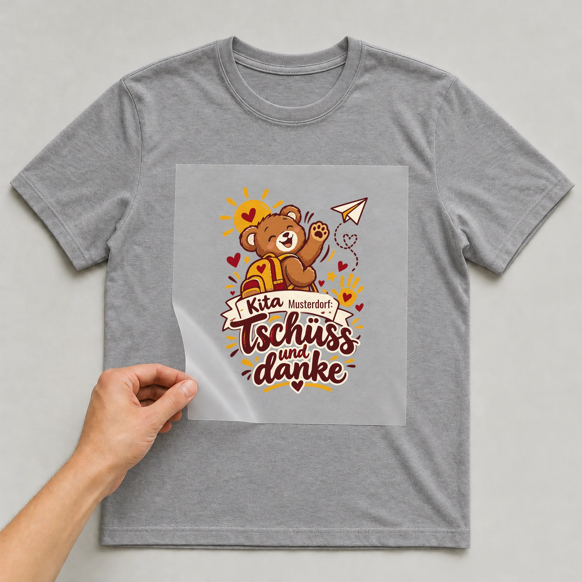 Bügelbild Kita: Tschüss und danke mit Ort auf grauem T-Shirt, Folie wird aufgelegt