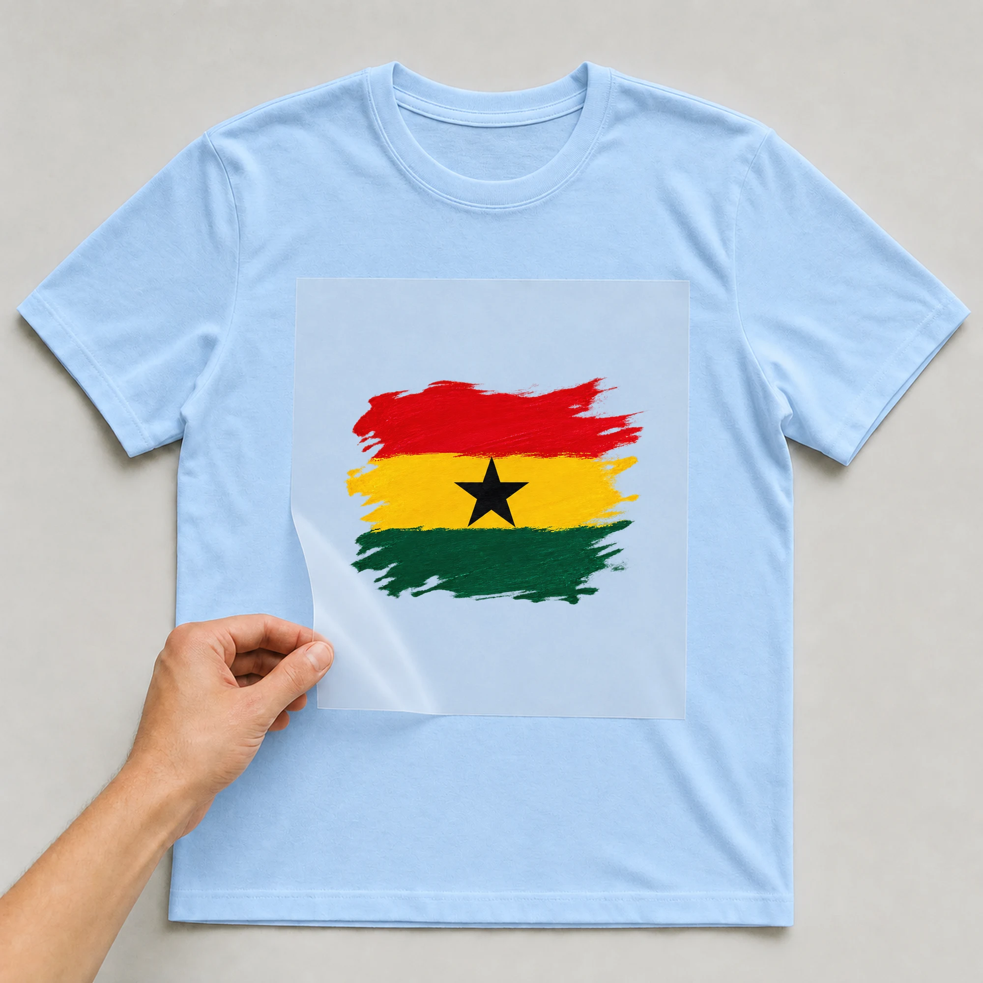 Bügelbild „Flagge Ghana im Pinselstrich“ auf babyblauem T-Shirt, Folie wird aufgelegt