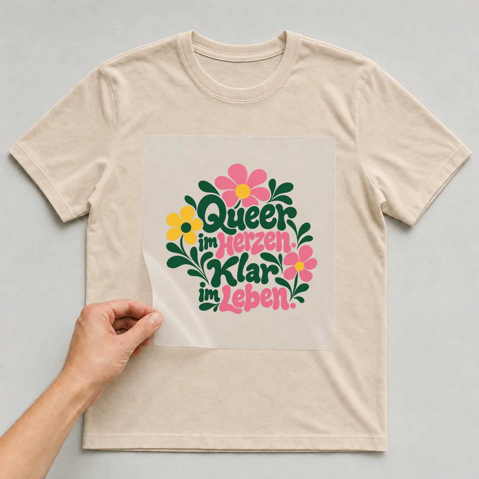 Bügelbild „Queer im Herzen. Klar im Leben.“ auf beigem T-Shirt, Folie wird aufgelegt