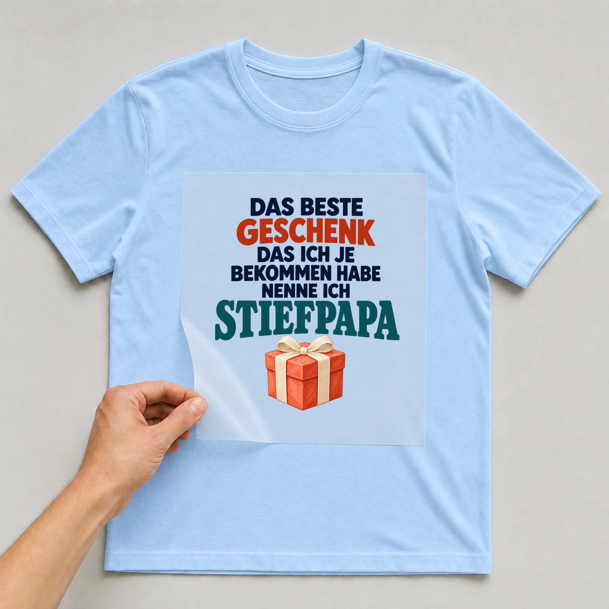 Bügelbild „Das beste Geschenk das ich je bekommen habe nenne ich Stiefpapa“ auf babyblauem T-Shirt, Folie wird aufgelegt