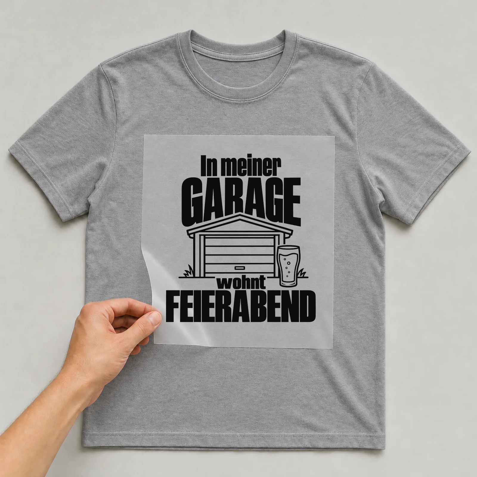 Bügelbild „In meiner Garage wohnt Feierabend“ auf grauem T-Shirt, Folie wird aufgelegt