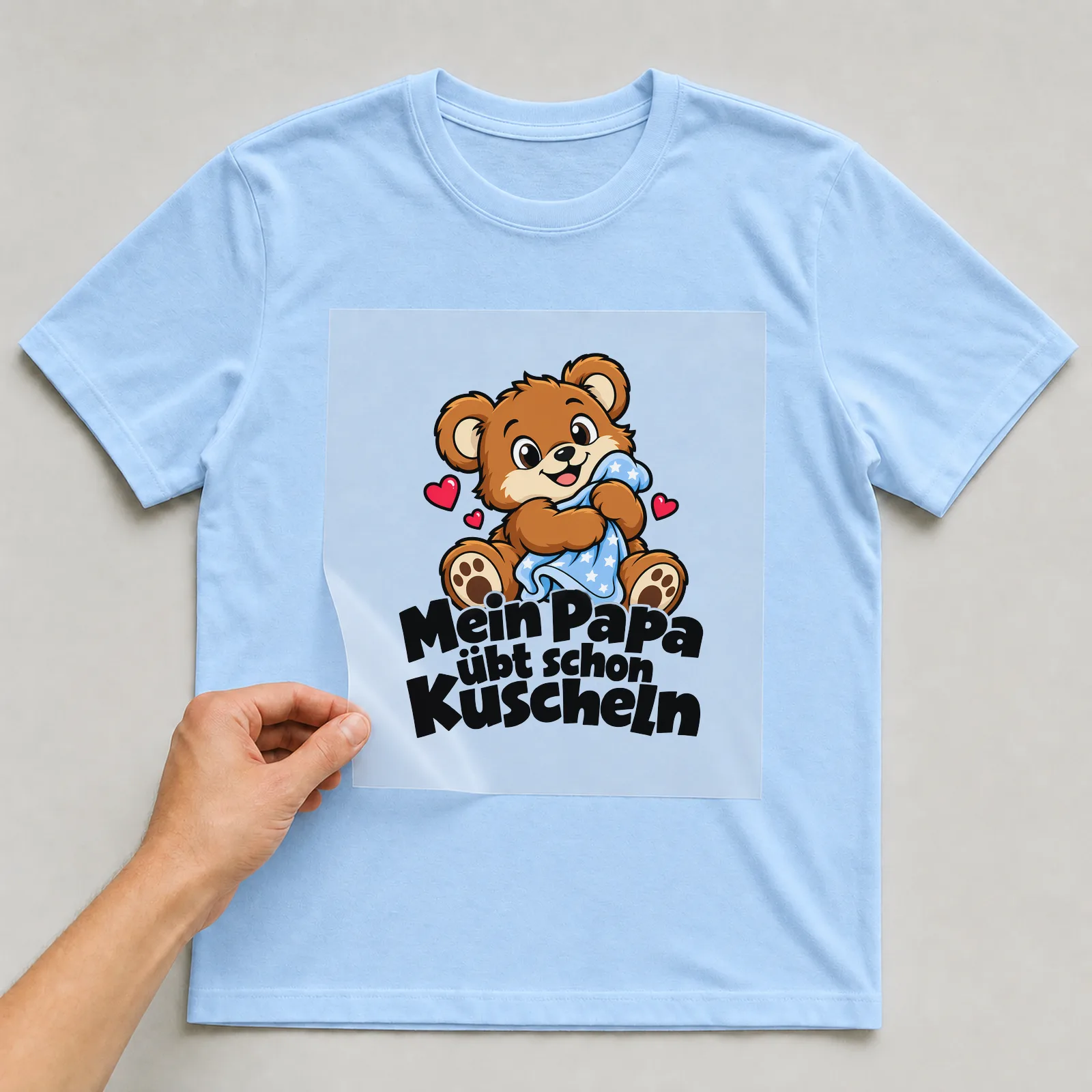 Bügelbild „Mein Papa übt schon Kuscheln“ auf hellblauem T-Shirt, Folie wird aufgelegt