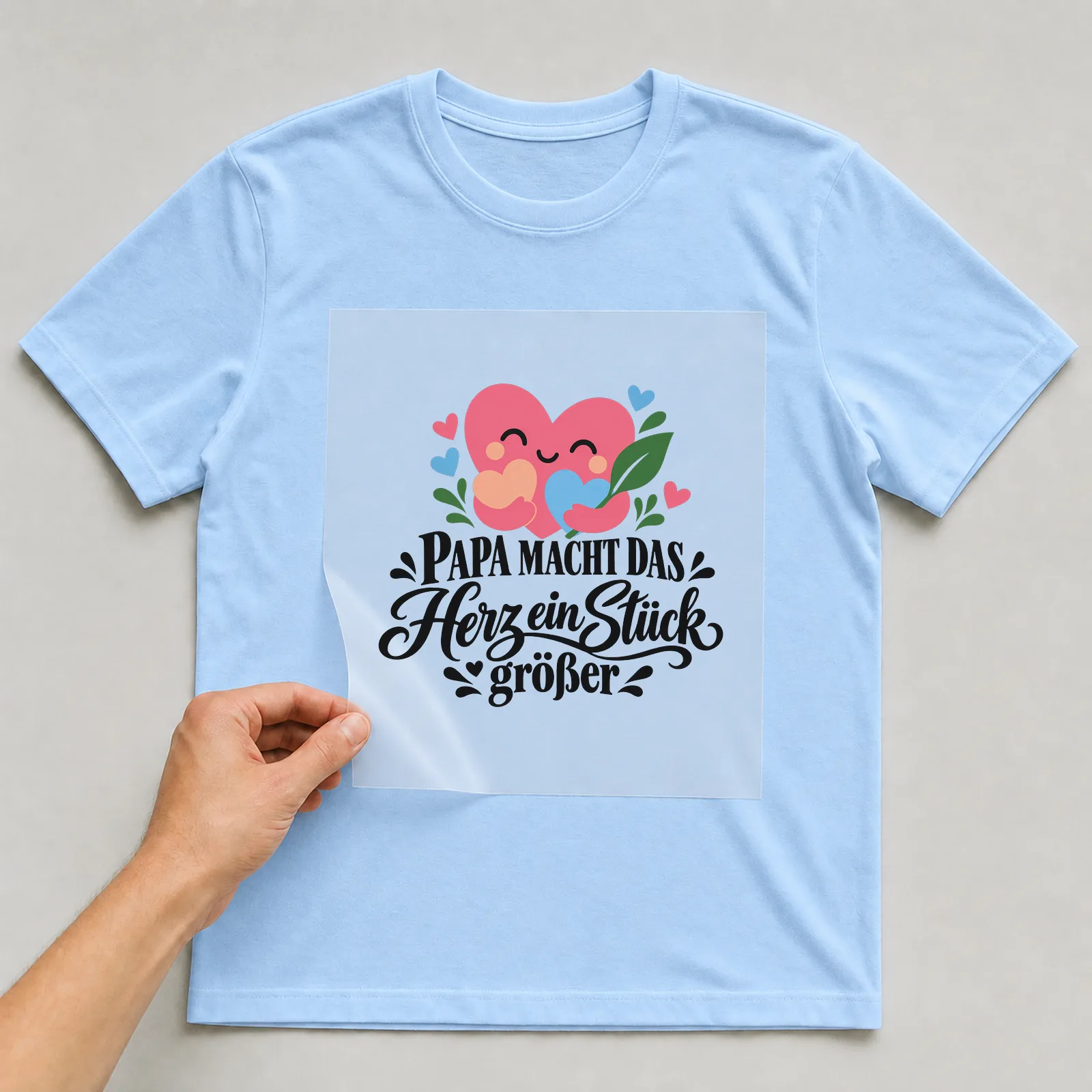 Bügelbild „PAPA MACHT DAS Herz ein Stück größer“ auf hellblauem T-Shirt, Folie wird aufgelegt