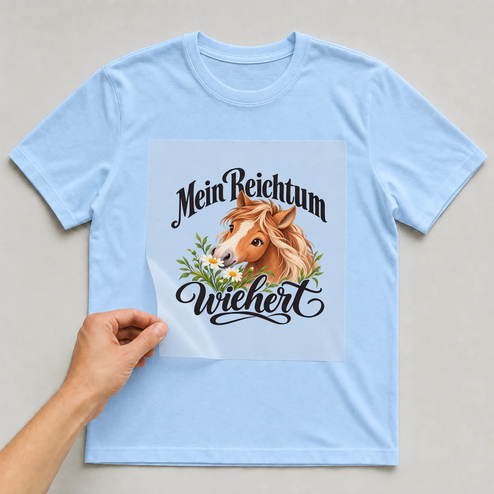 Bügelbild „Mein Reichtum wiehert“ auf hellblauem T-Shirt, Folie wird aufgelegt