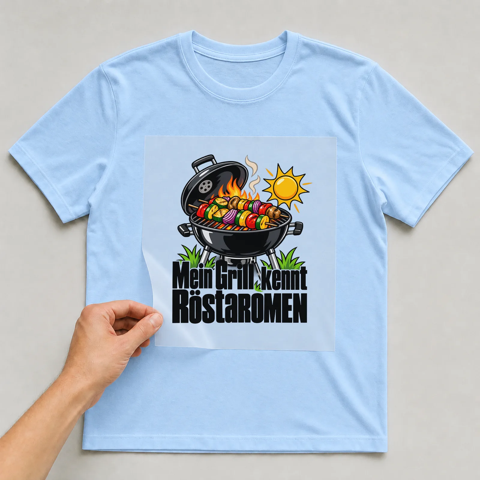 Bügelbild „Mein Grill kennt Röstaromen“ auf hellblauem T-Shirt, Folie wird aufgelegt