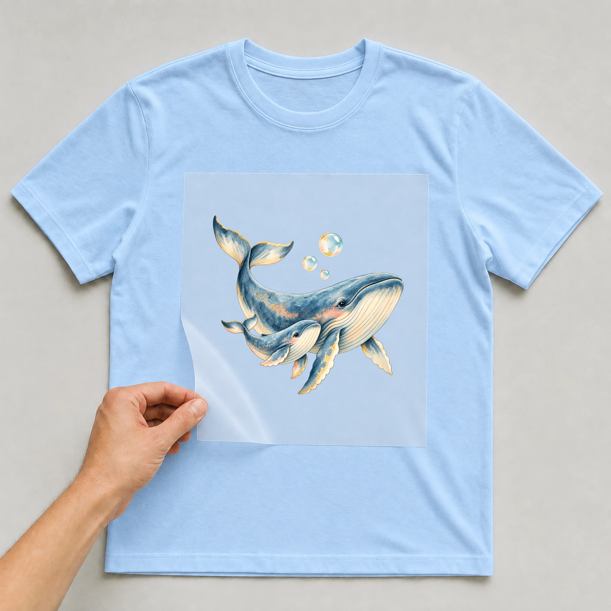 Bügelbild „Wal voller Meeresliebe“ auf babyblauem T-Shirt, Folie wird aufgelegt