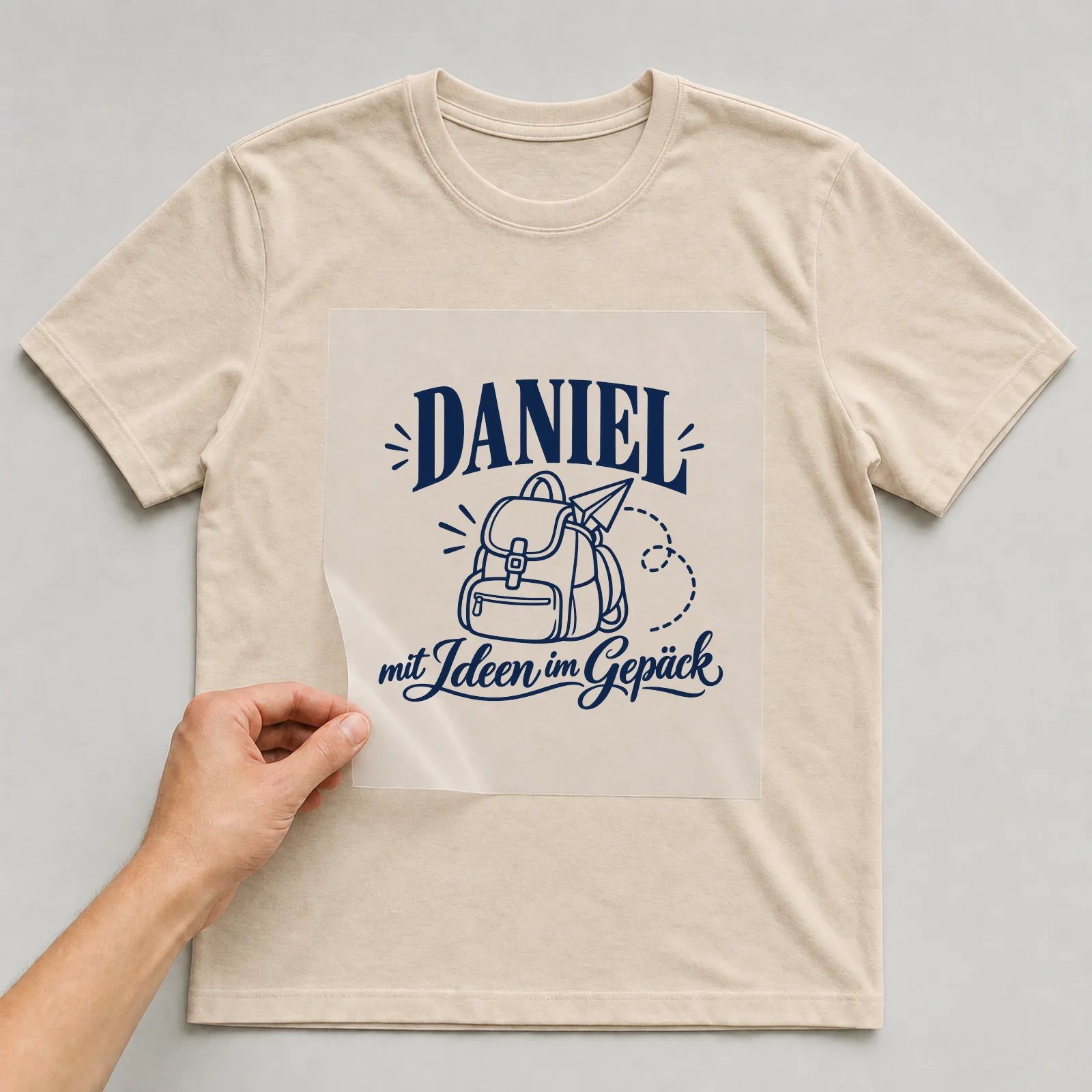 Bügelbild „Daniel mit Ideen im Gepäck“ auf beigem T-Shirt, Folie wird aufgelegt
