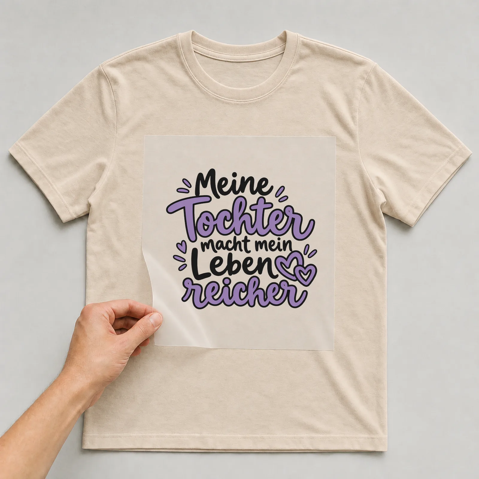Bügelbild „Meine Tochter macht mein Leben reicher“ auf beigem T-Shirt, Folie wird aufgelegt