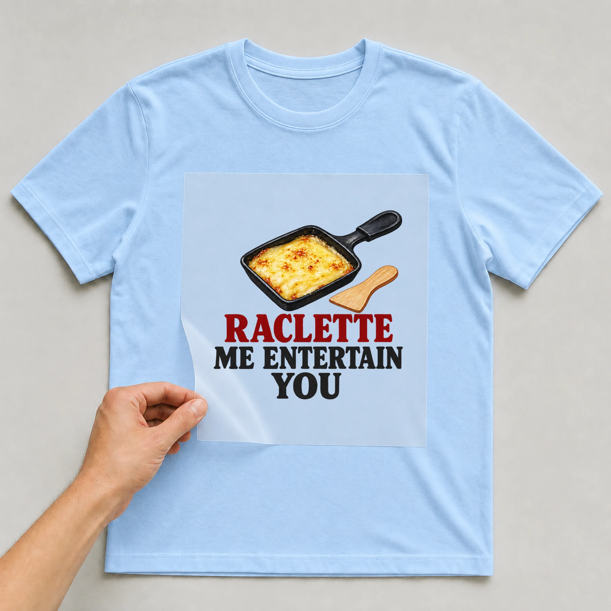 Bügelbild „Raclette Me Entertain You“ auf babyblauem T-Shirt, Folie wird aufgelegt
