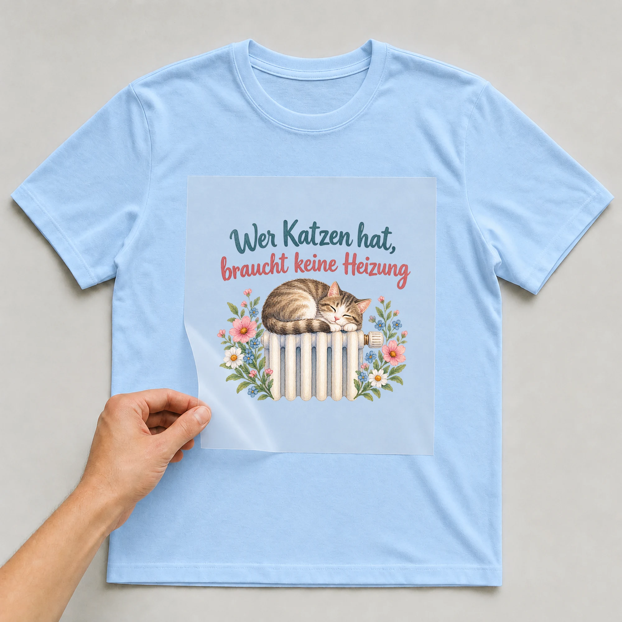 Bügelbild „Wer Katzen hat, braucht keine Heizung“ auf babyblauem T-Shirt, Folie wird aufgelegt