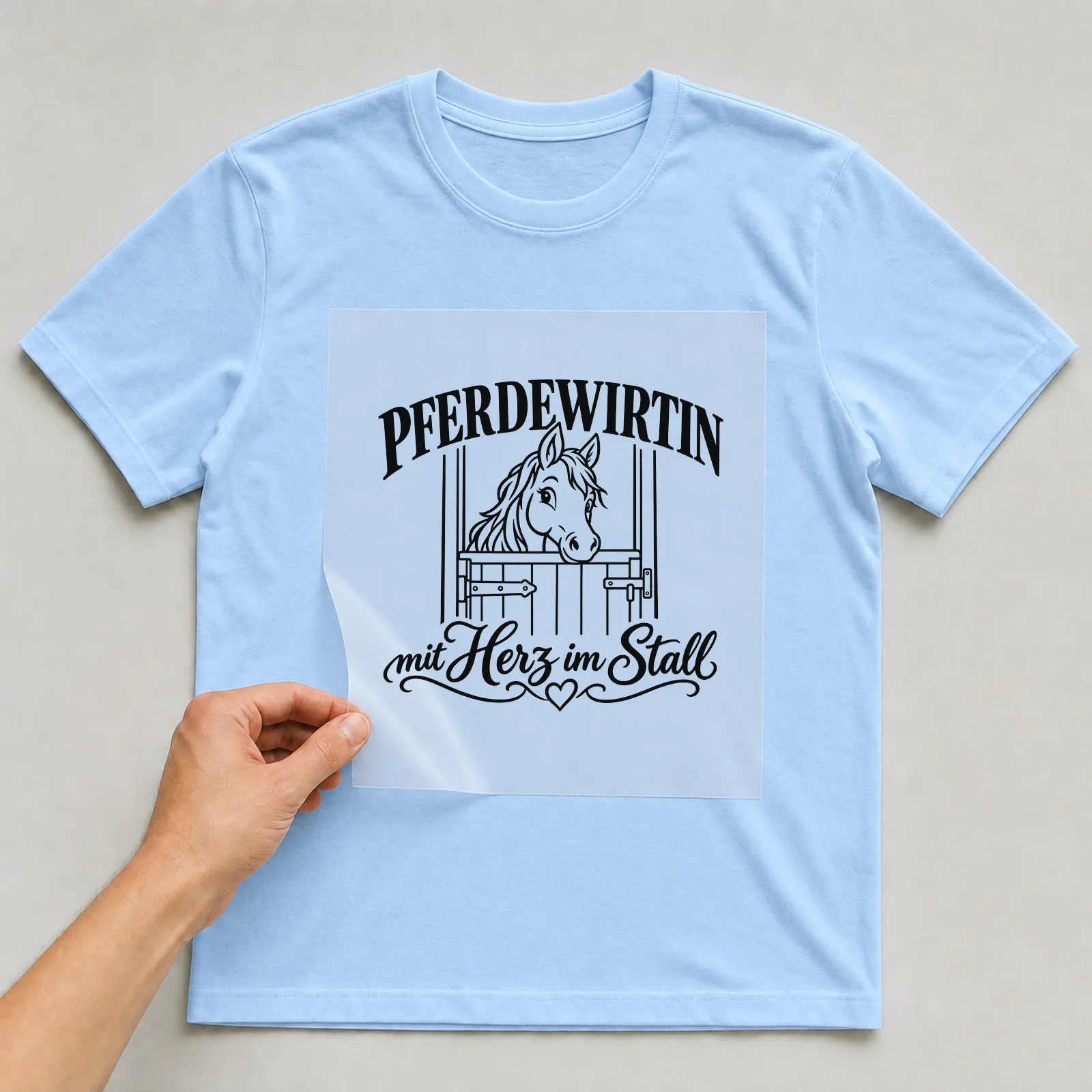 Bügelbild „Pferdewirtin mit Herz im Stall“ auf hellblauem T-Shirt, Folie wird aufgelegt