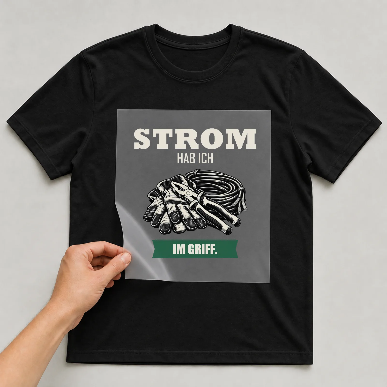 Bügelbild „Strom hab ich im Griff“ auf schwarzem T-Shirt, Folie wird aufgelegt