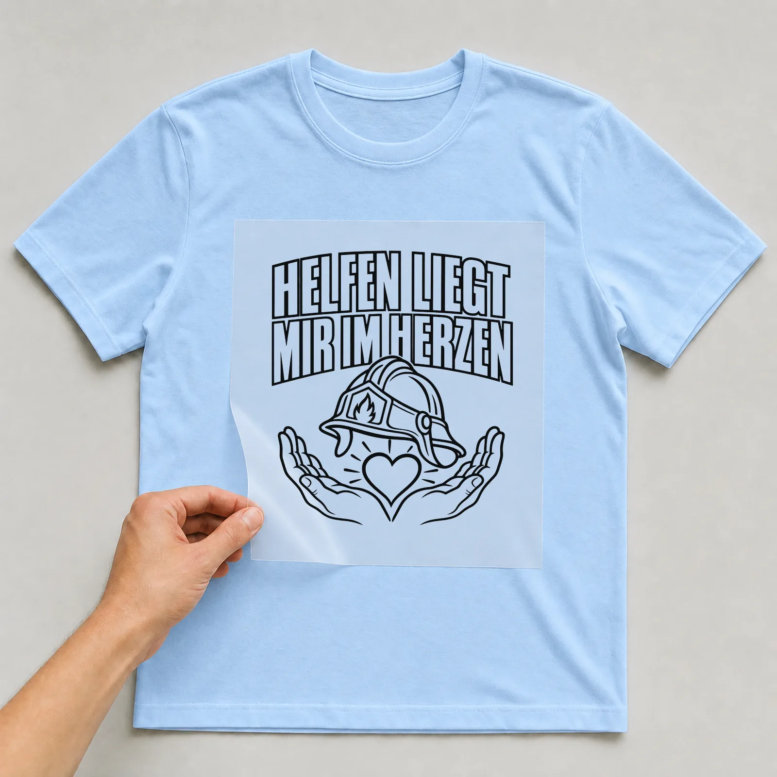 Bügelbild „Helfen liegt mir im Herzen“ auf hellblauem T-Shirt, Folie wird aufgelegt