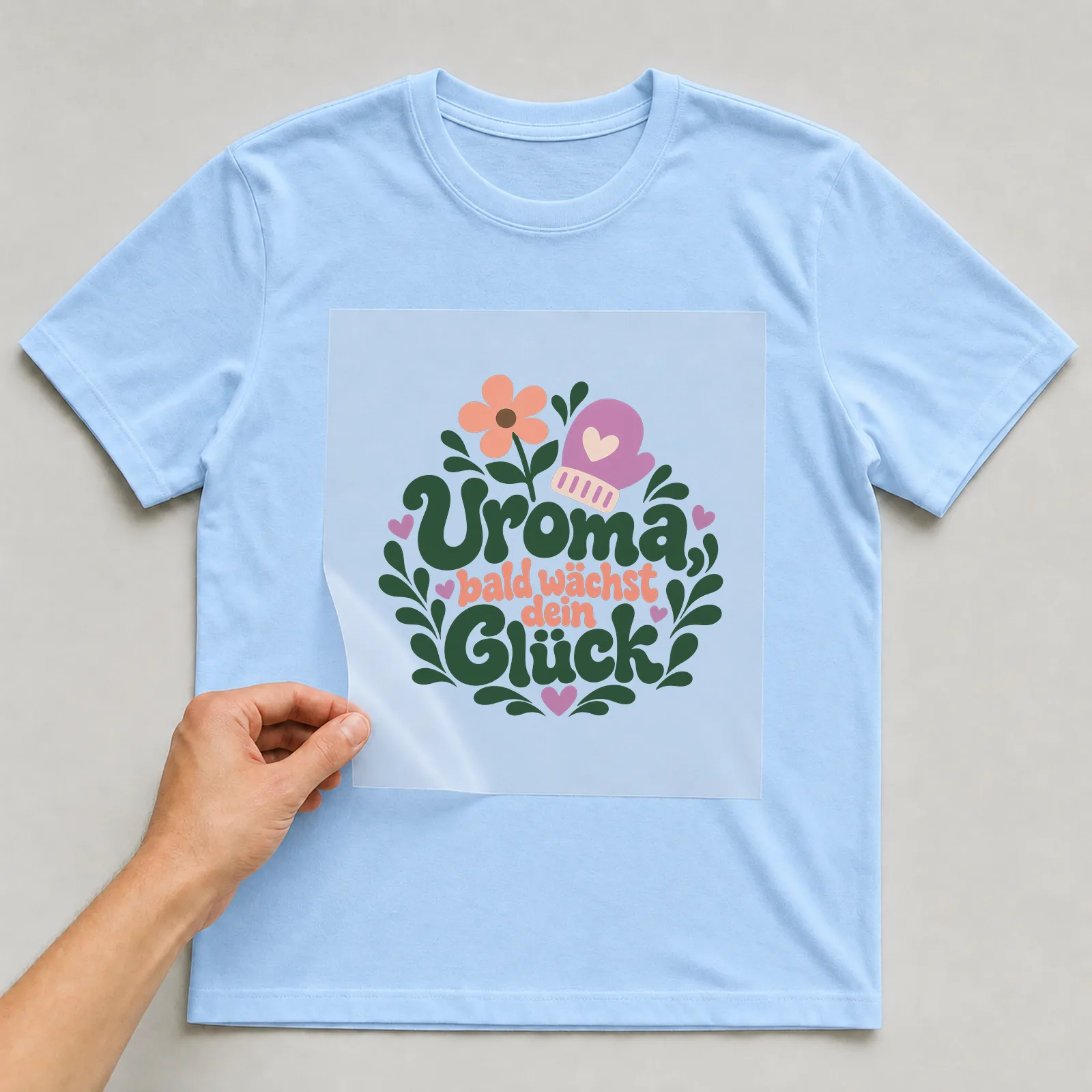 Bügelbild „Uroma, bald wächst dein Glück“ auf hellblauem T-Shirt, Folie wird aufgelegt