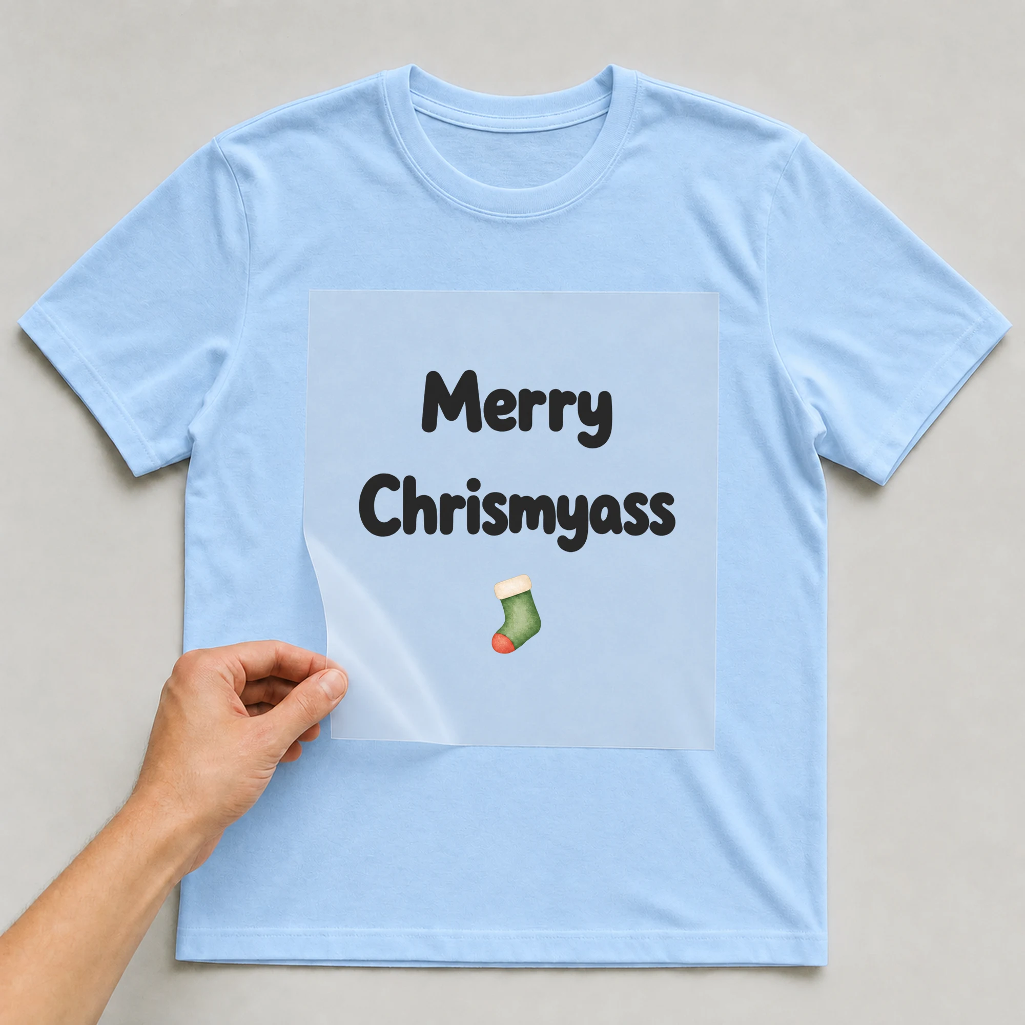 Bügelbild „Merry Chrismyass“ auf babyblauem T-Shirt, Folie wird aufgelegt