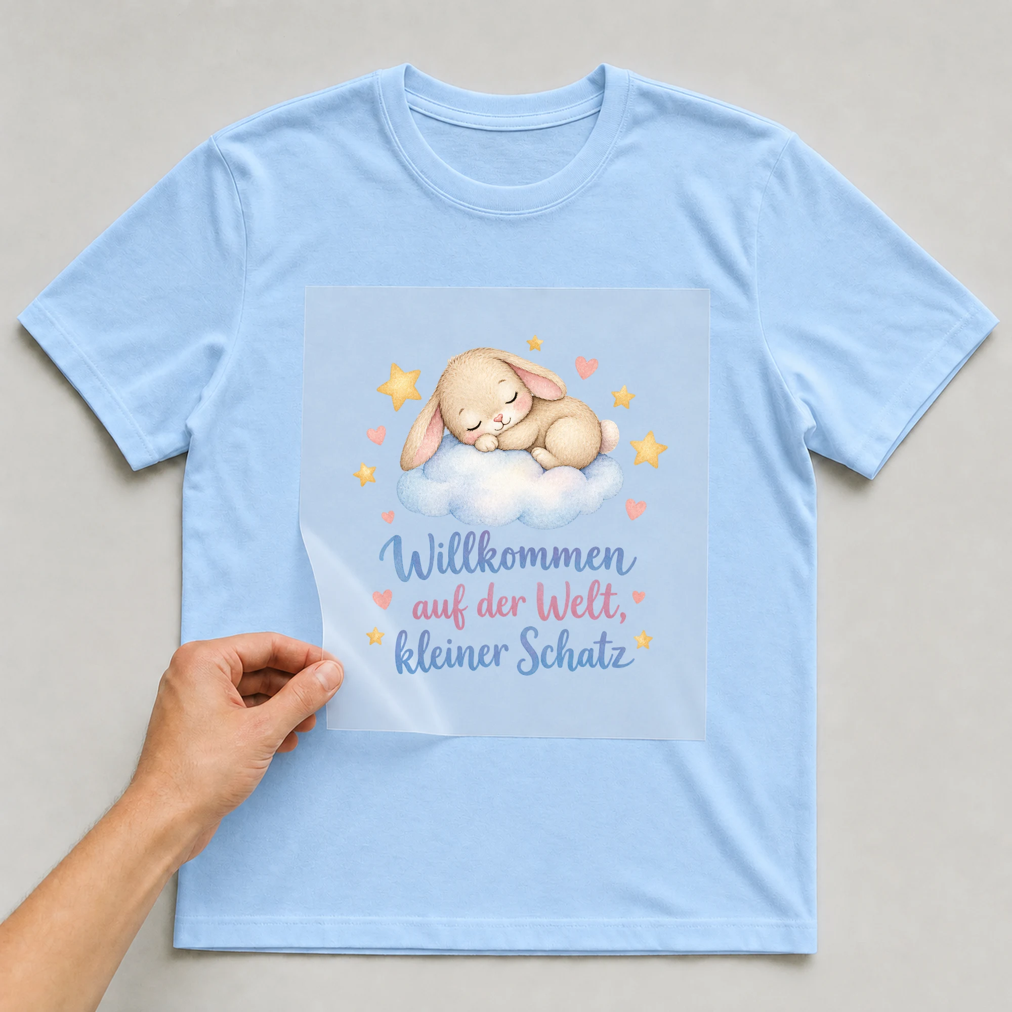 Bügelbild „Willkommen auf der Welt, kleiner Schatz“ auf babyblauem T-Shirt, Folie wird aufgelegt